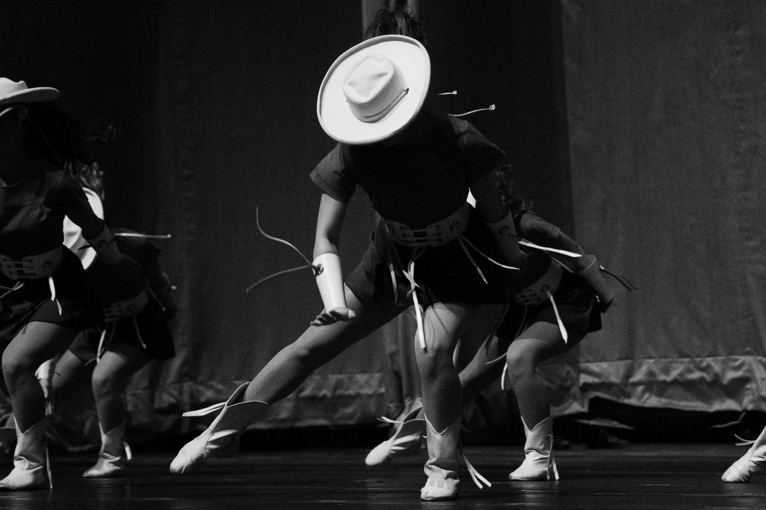 Black and white photo of dancers performing on stage in matching outfits and hats during a choreographed routine.