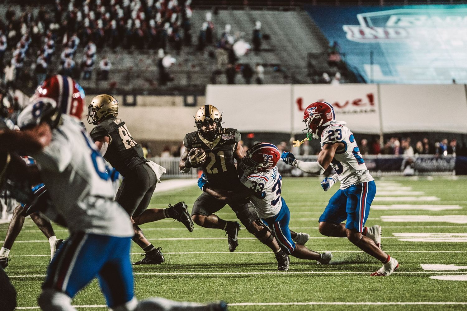 Football players in blue and dark uniforms compete during a night game at a stadium.