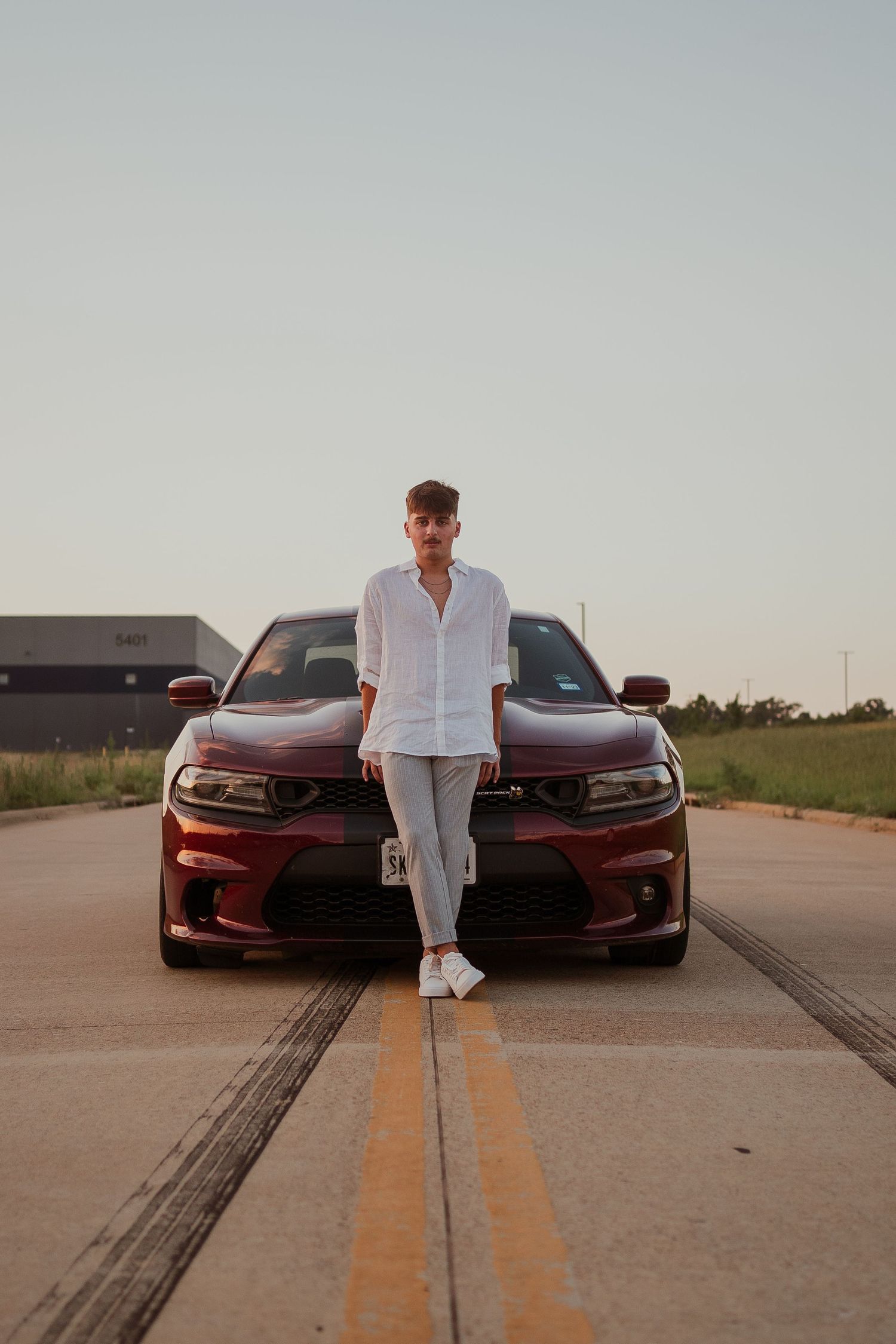 Person in all white outfit poses between two red cars on empty parking lot at sunset.