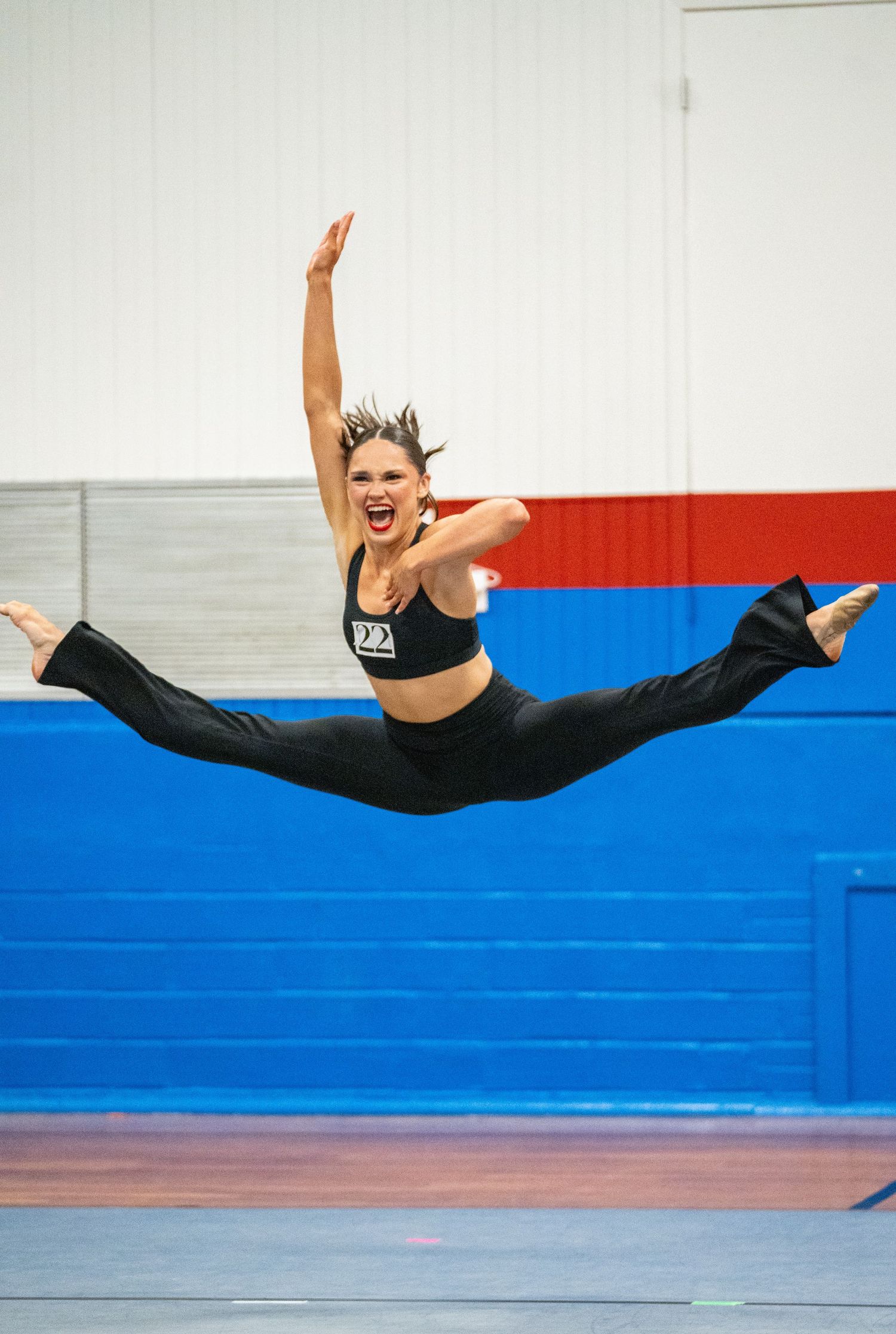 Female dancer performs a stunning mid-air split leap in a black crop top and pants inside a gymnasium.