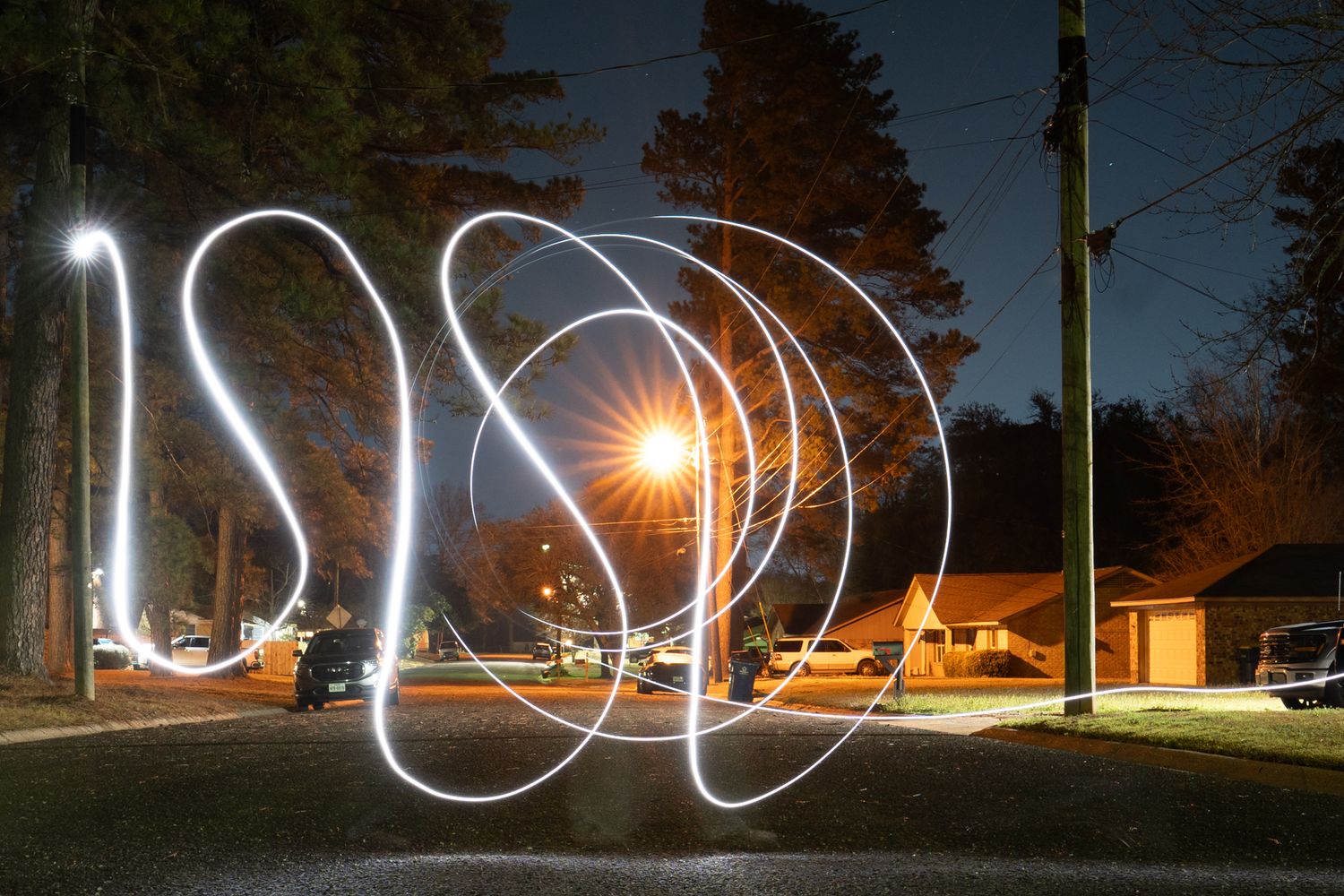 Long exposure photograph captures light trails creating abstract patterns against a dark night sky in a park setting.