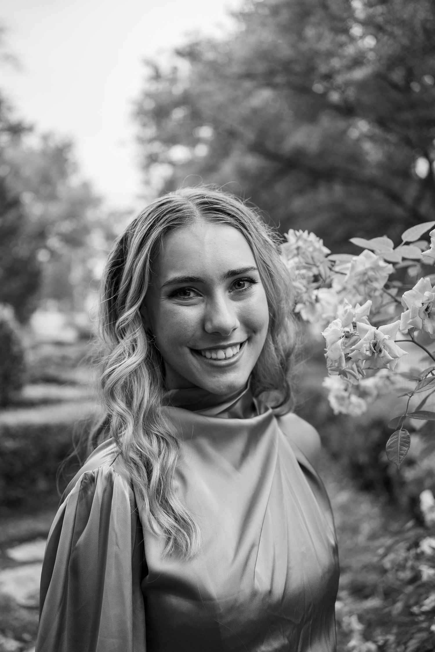 Black and white portrait of someone in elegant dress posed among flowering bushes.