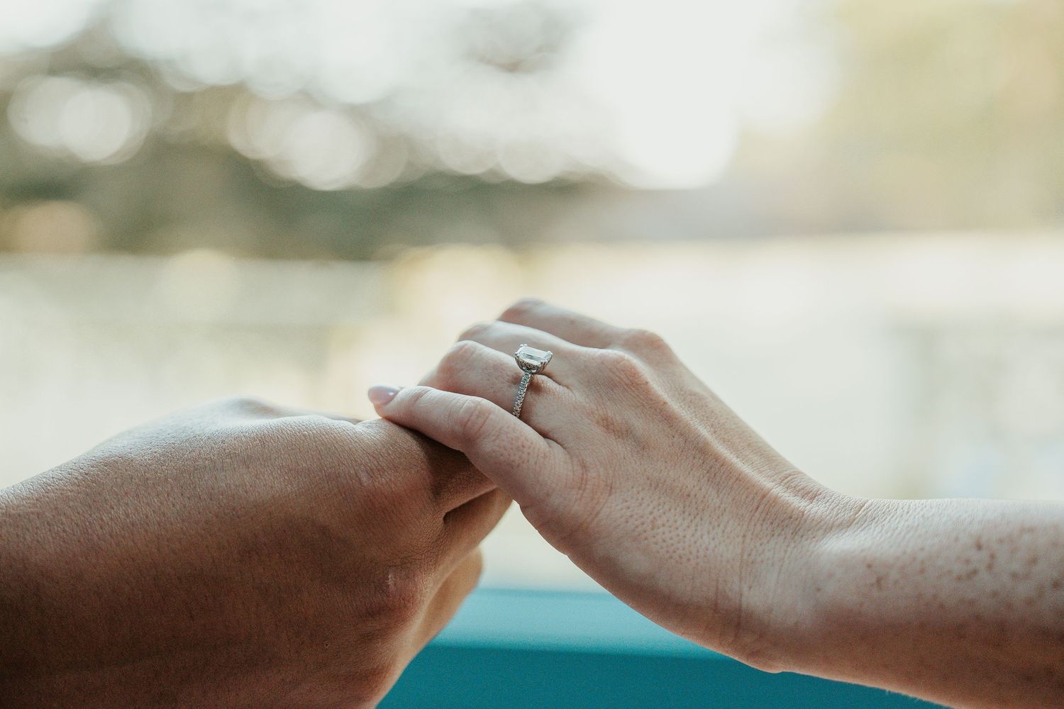Close-up of hands with engagement ring holding each other against blurred background.
