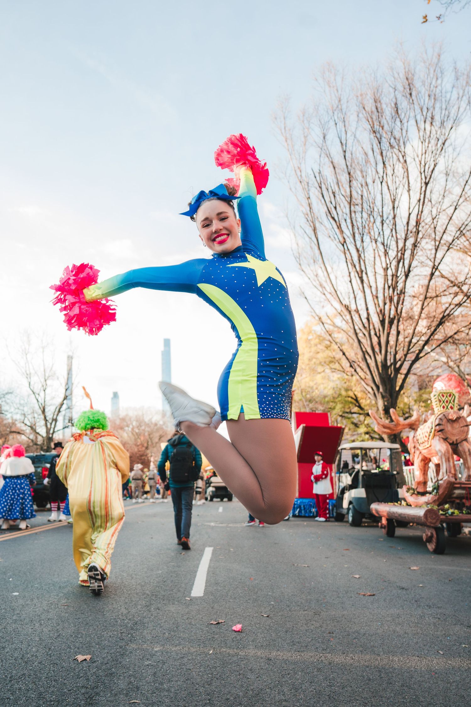 A cheerleader in blue performs dynamic jumps and poses during a holiday parade on a street with spectators.