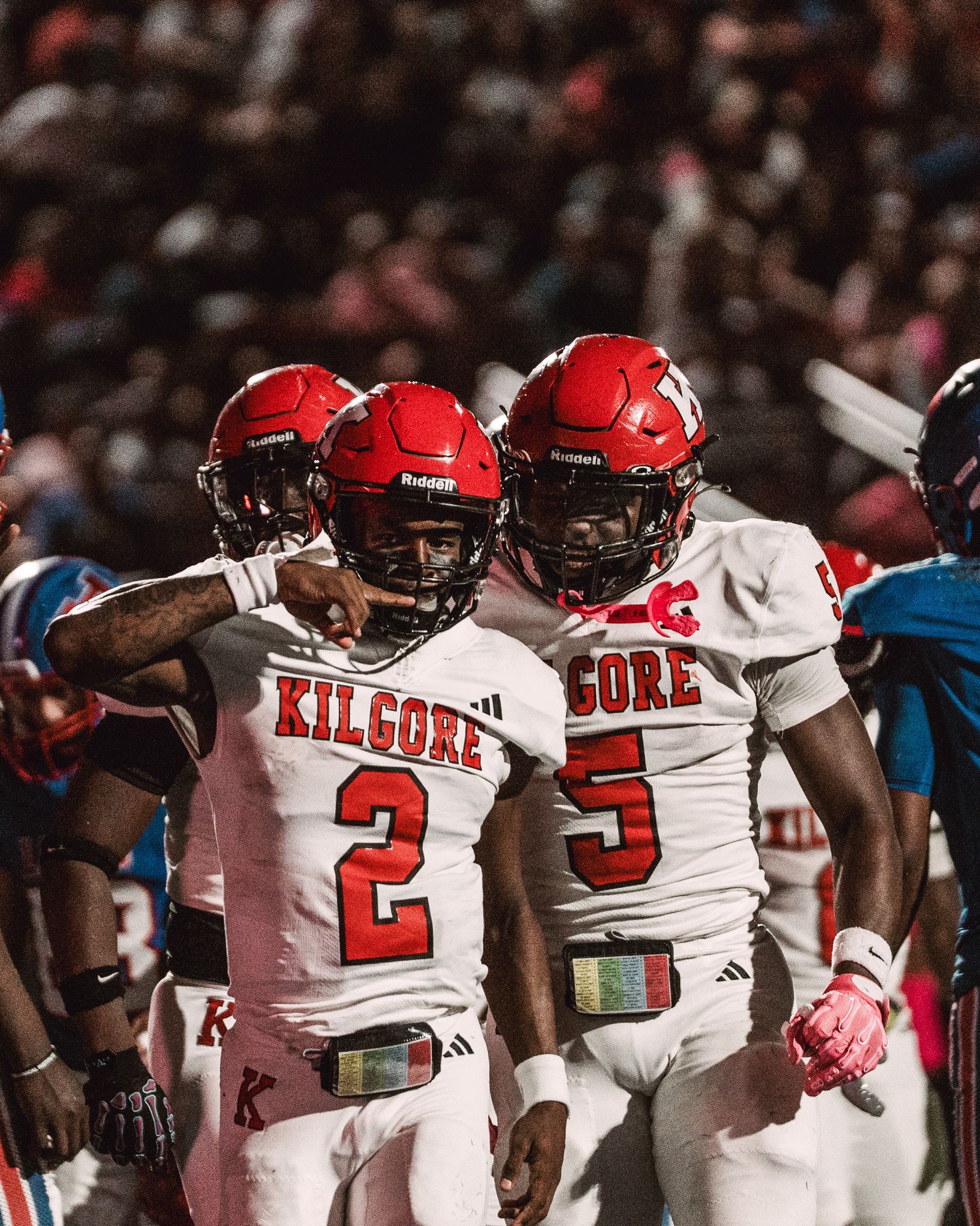 Kilgore football players in red helmets and white jerseys celebrate on the field during a night game.