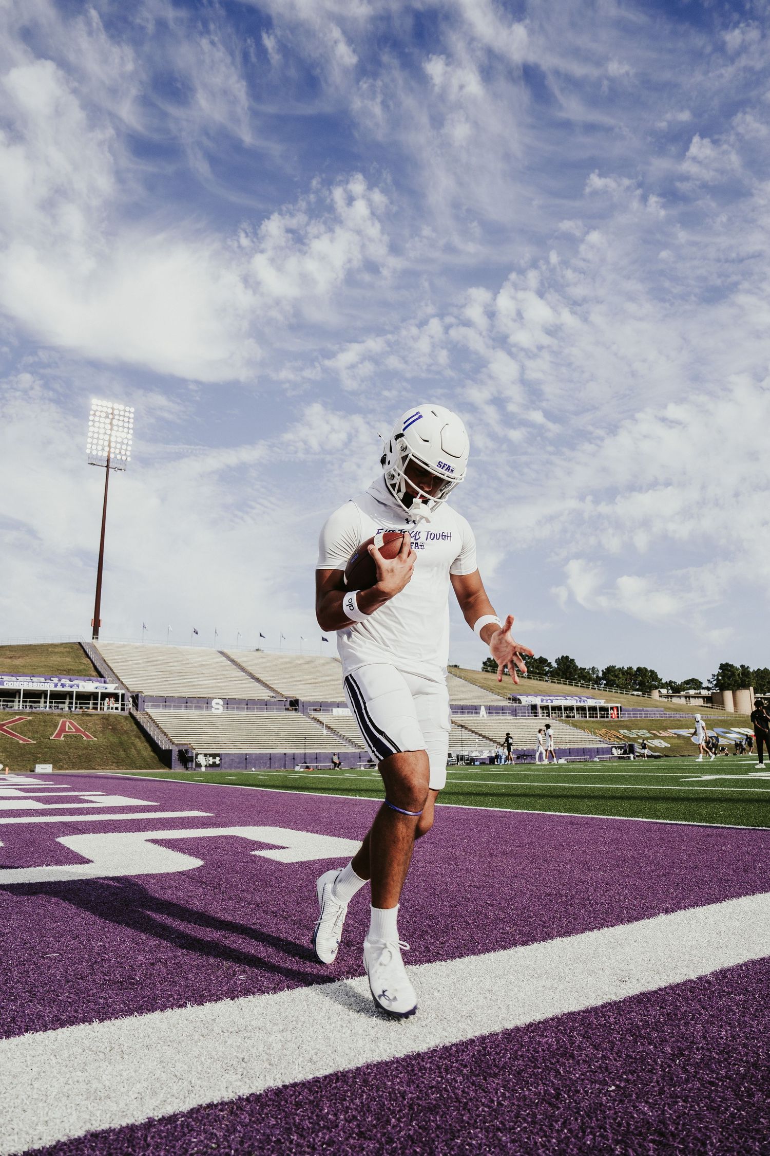 Football player in white uniform practices stance and form drills on purple and white turf field under cloudy sky.