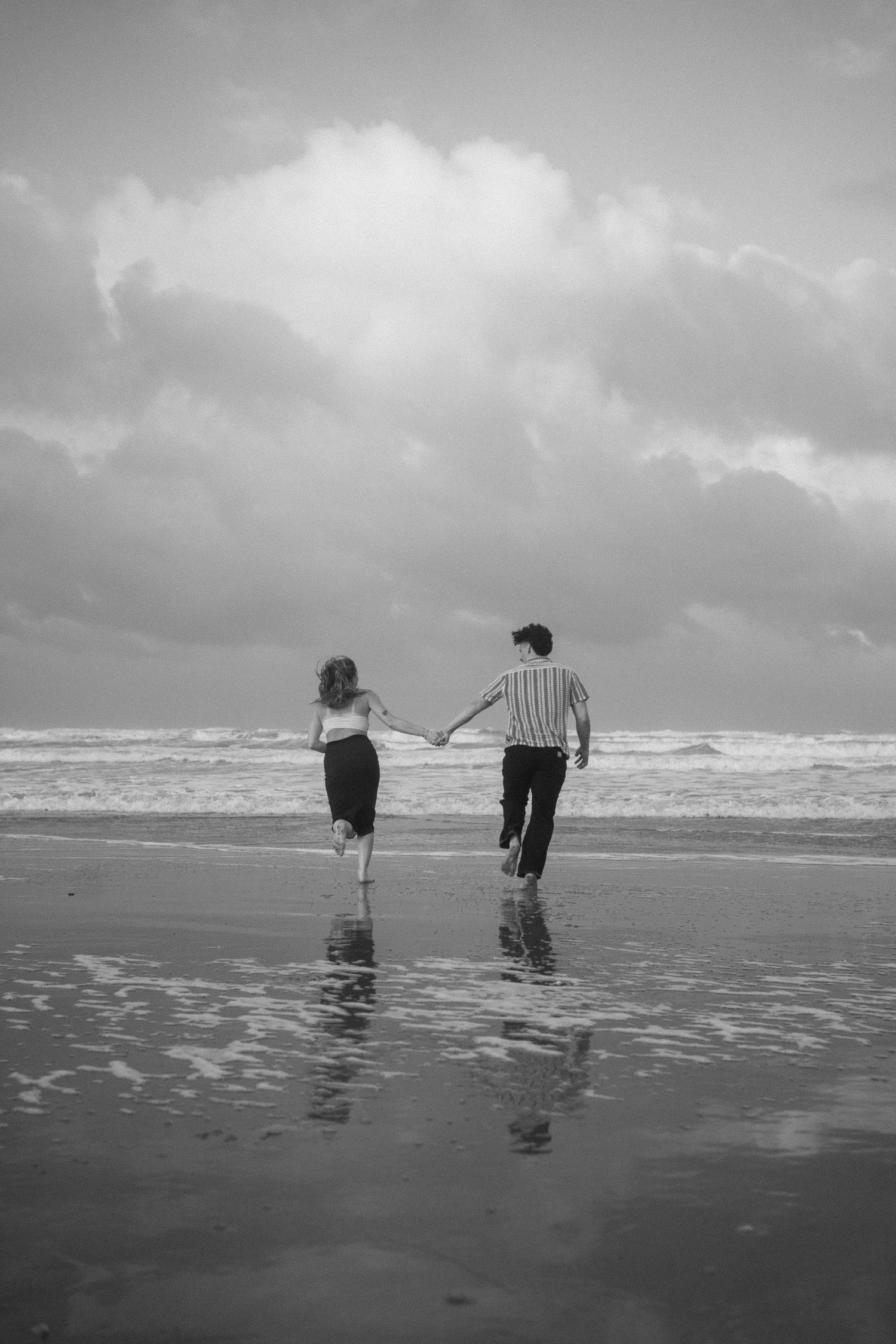 Couple walks hand in hand along beach shoreline in black and white photograph.
