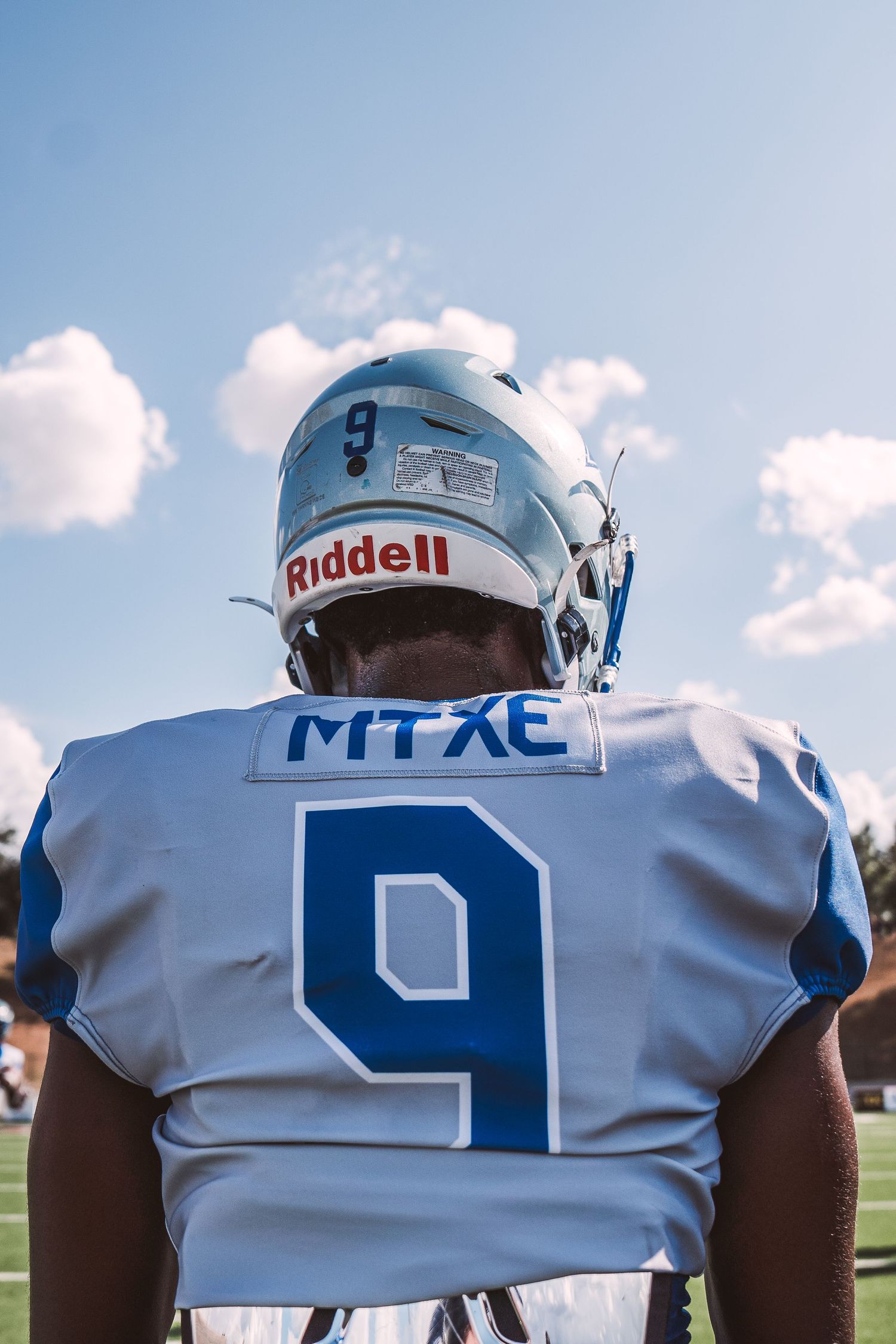 Row of football players wearing white jerseys with blue numbers viewed from behind.