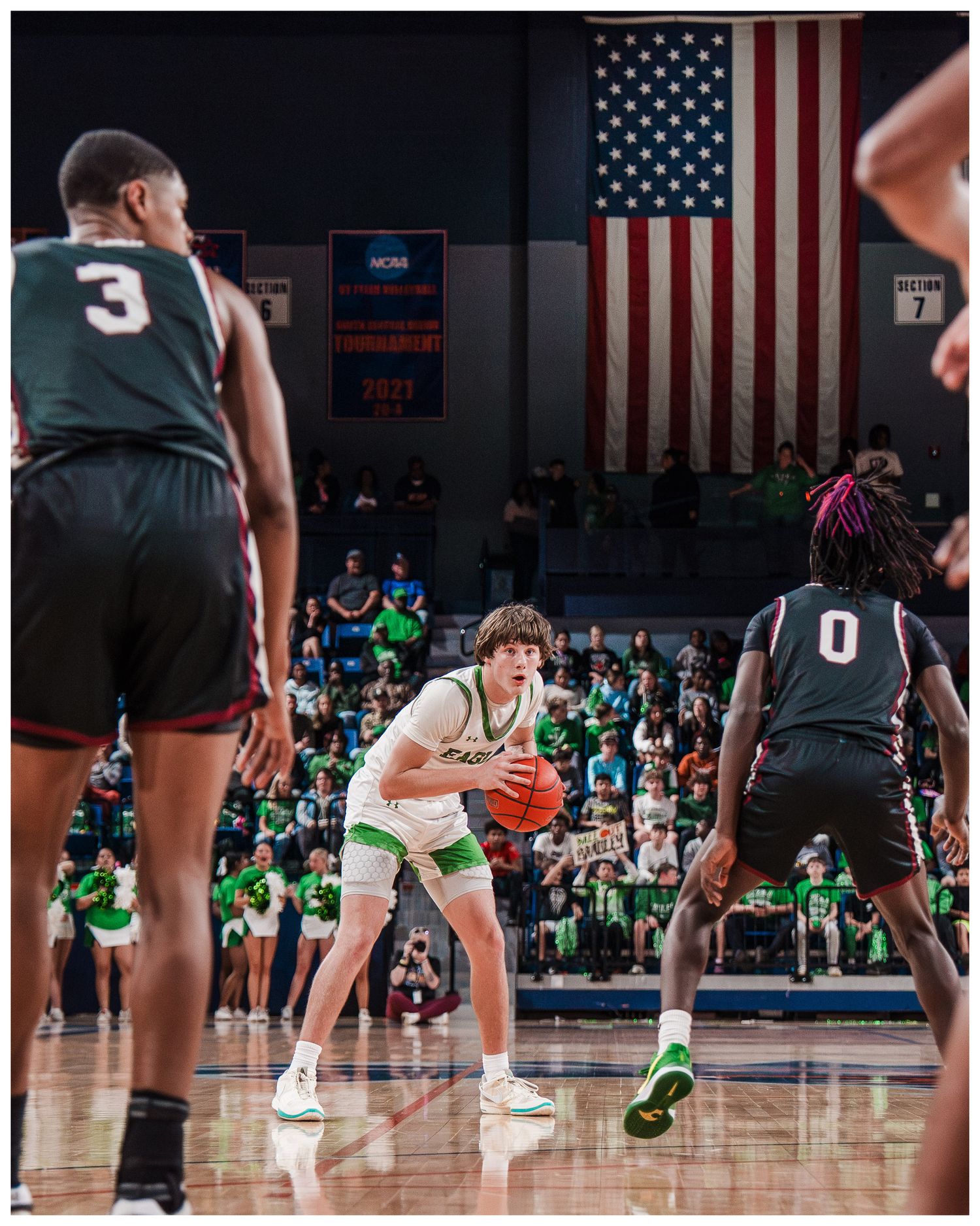 Basketball game in progress with American flag backdrop in indoor gymnasium.