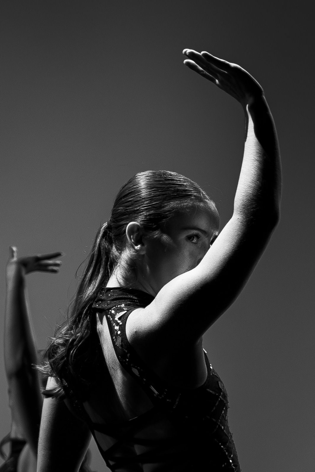 Black and white photo of a female dancer with arm raised gracefully, wearing a sequined costume, captured from behind.