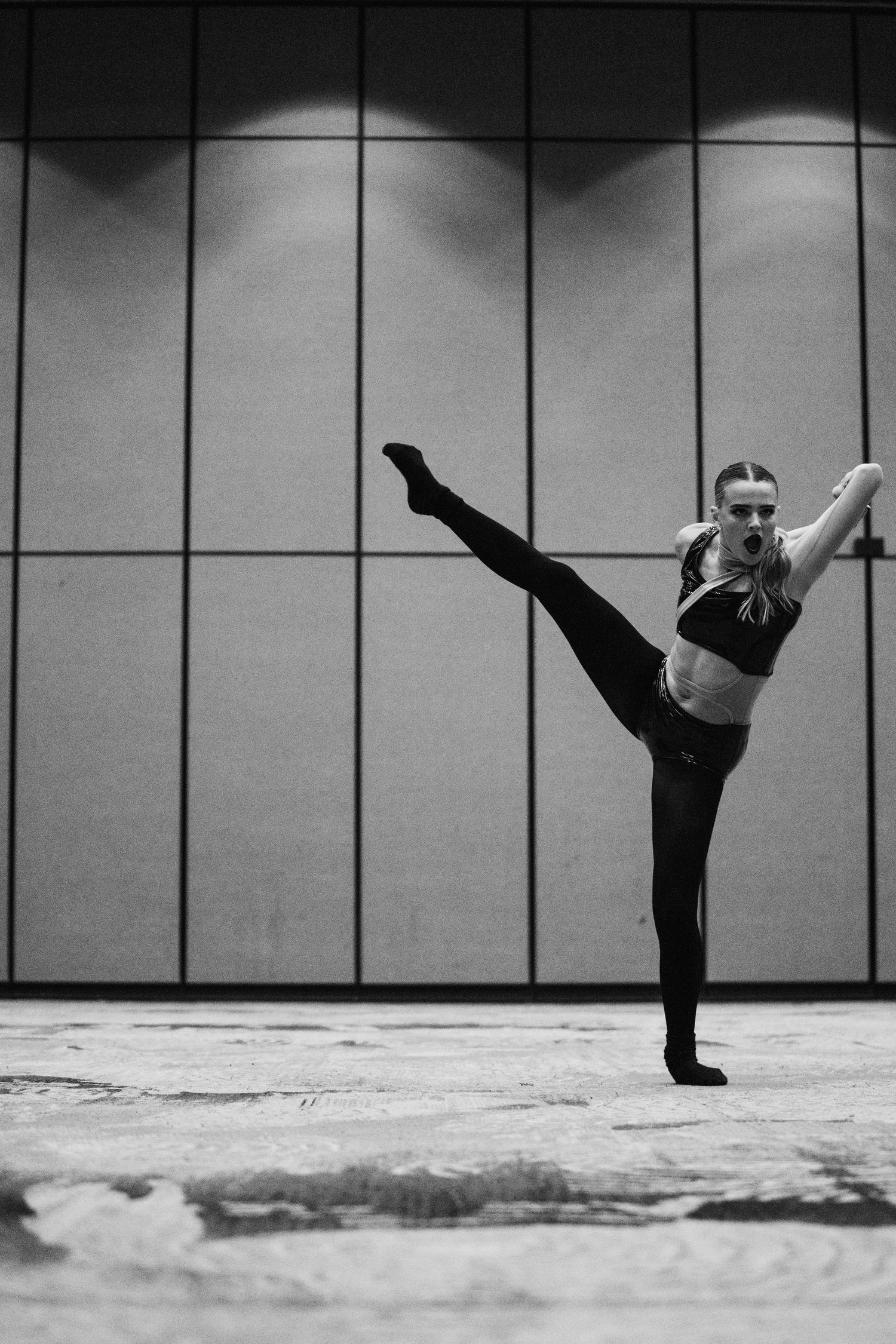 Black and white photo of a female dancer performing a high kick against a geometric wall backdrop.