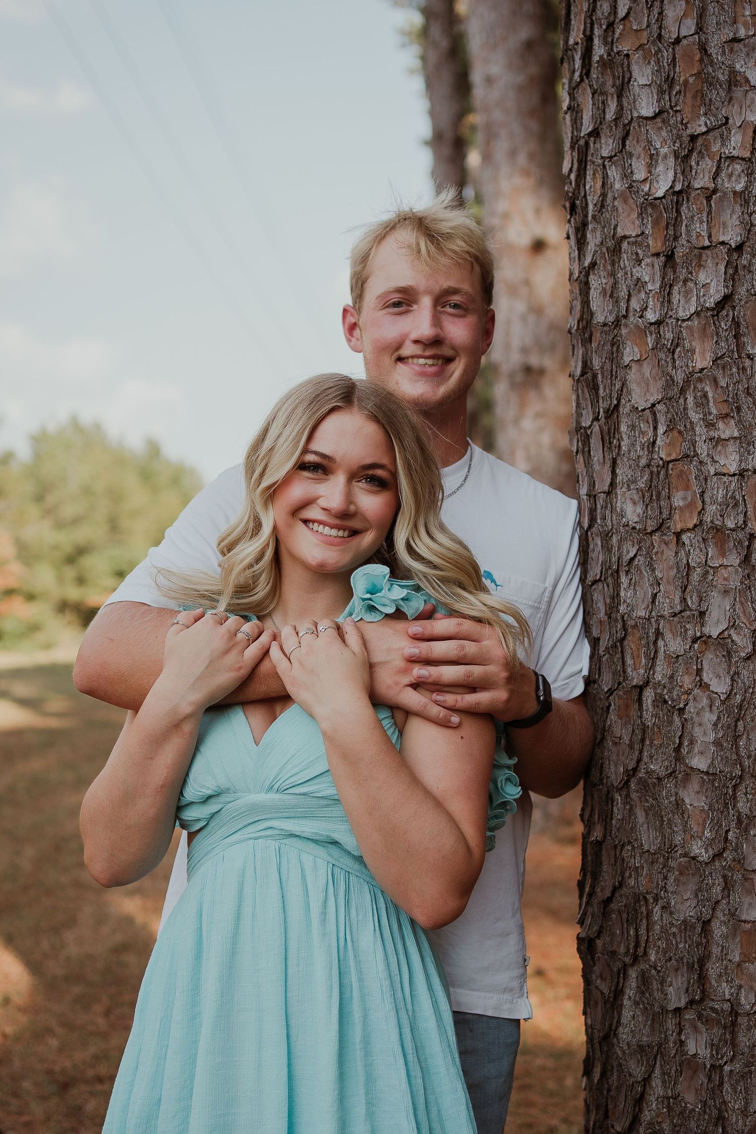 Couple standing by tree trunk outdoors, woman wearing turquoise dress.