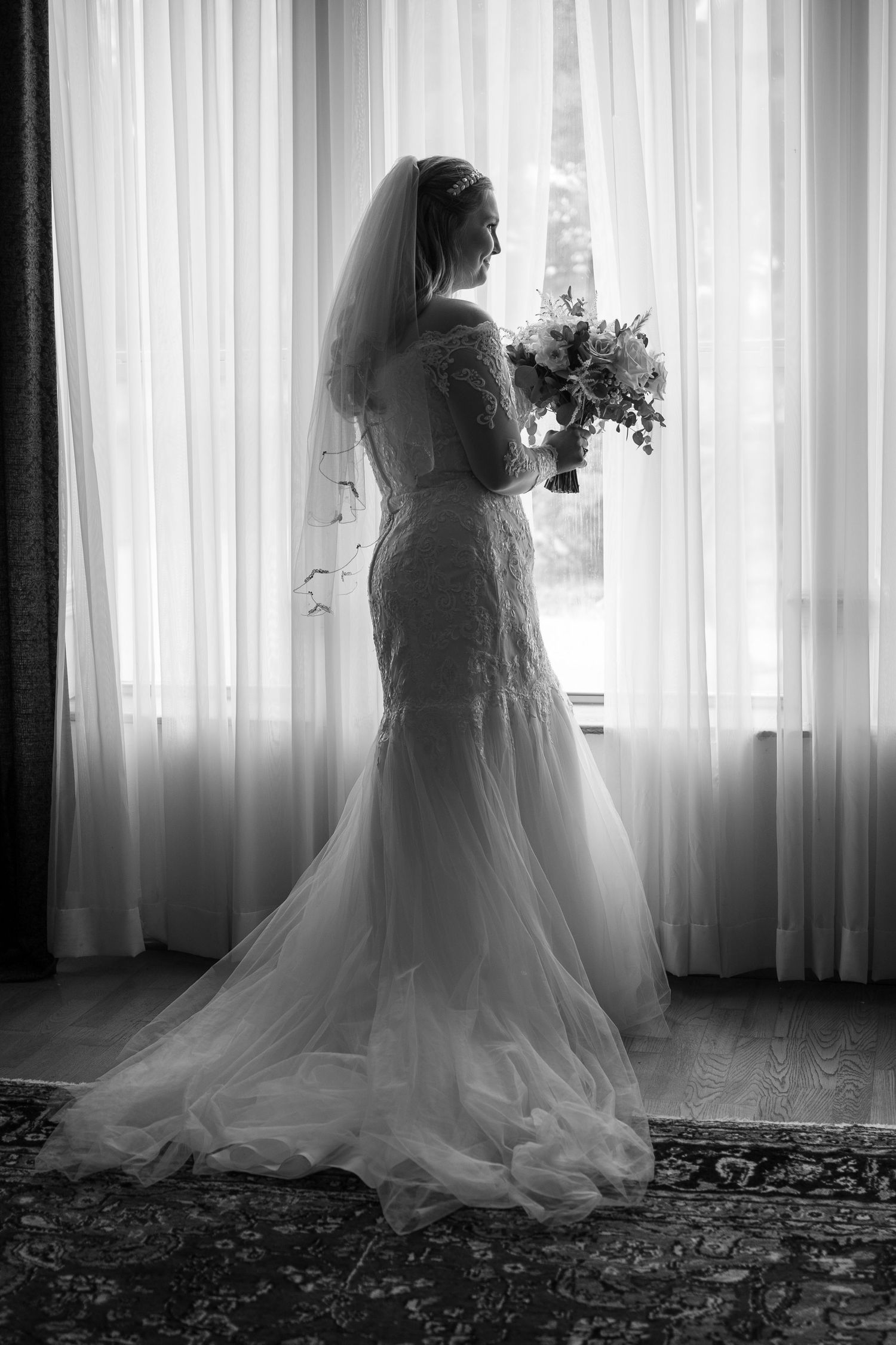 Silhouette of bride in lace gown holding bouquet while standing by window with sheer curtains.