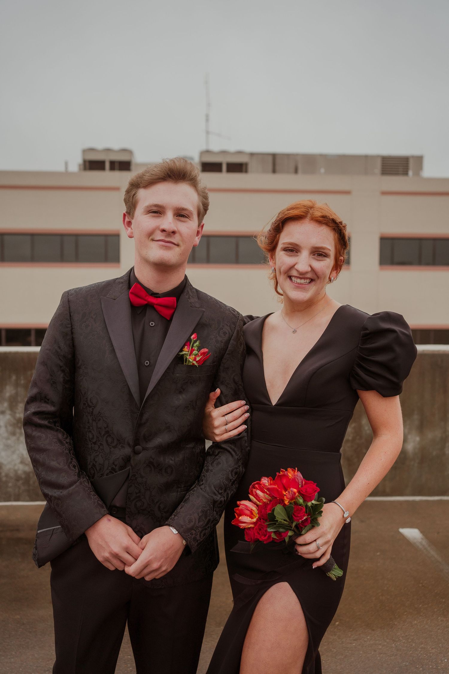 Formal couple posing outdoors in black attire with red roses and bow tie for special occasion.