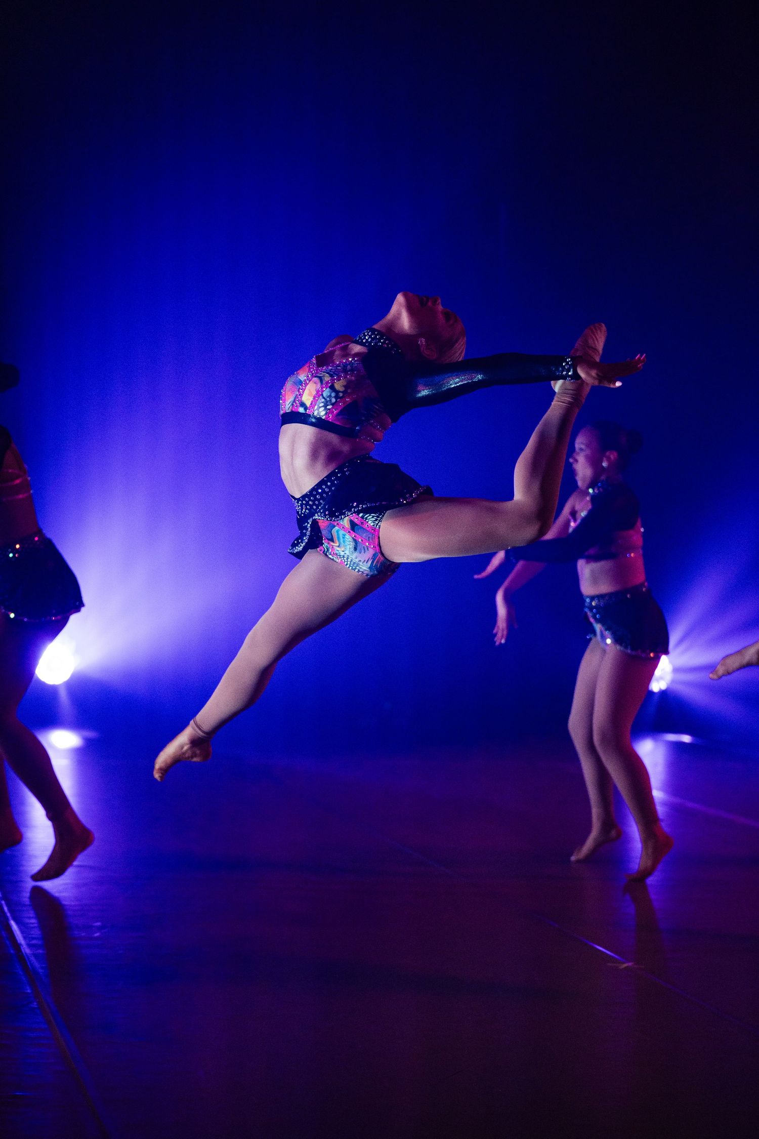 Dancers perform on a blue-lit stage, one leaping high in colorful costume during a dynamic dance recital.