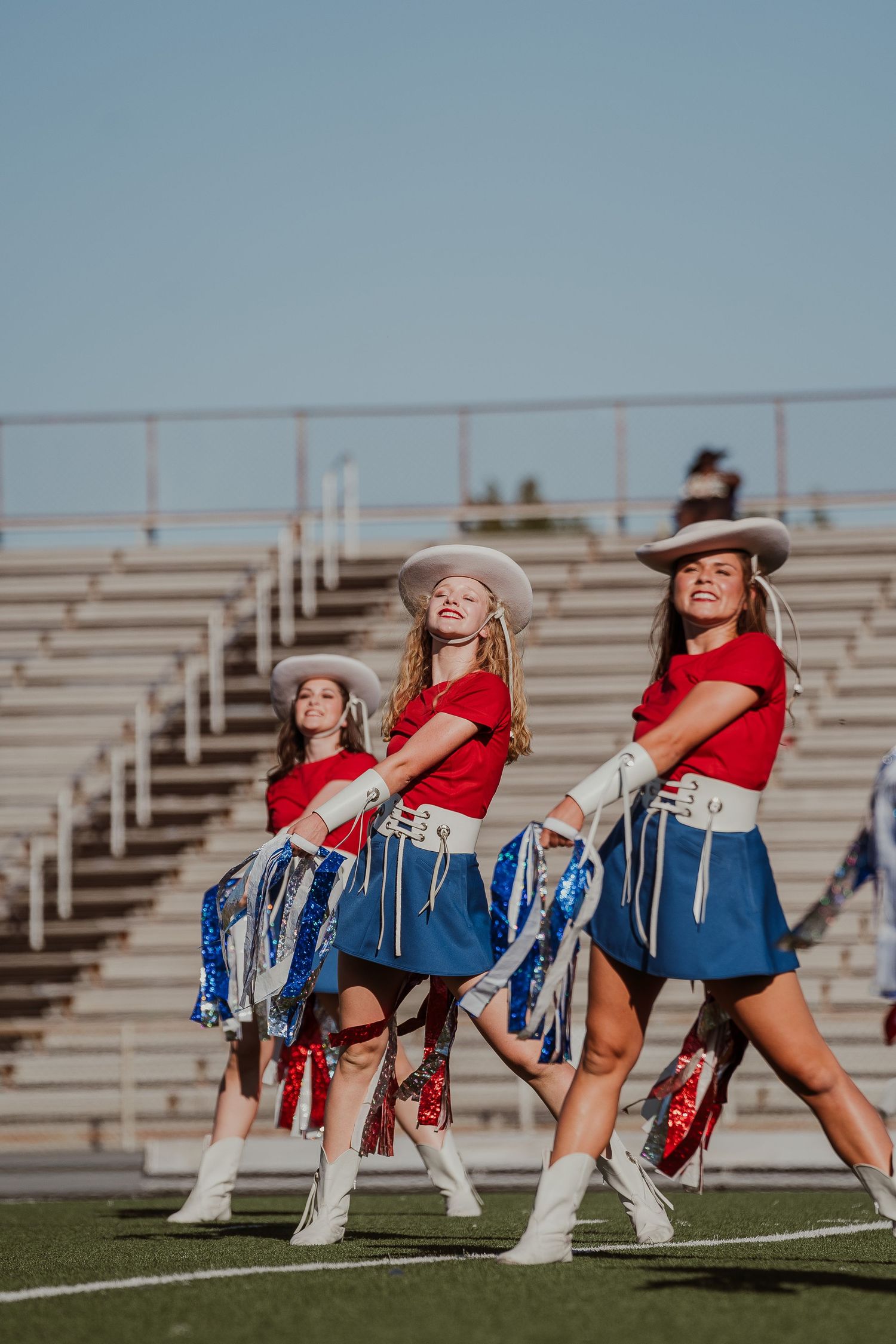 Dance team members in red tops and blue skirts performing synchronized routine on football field.