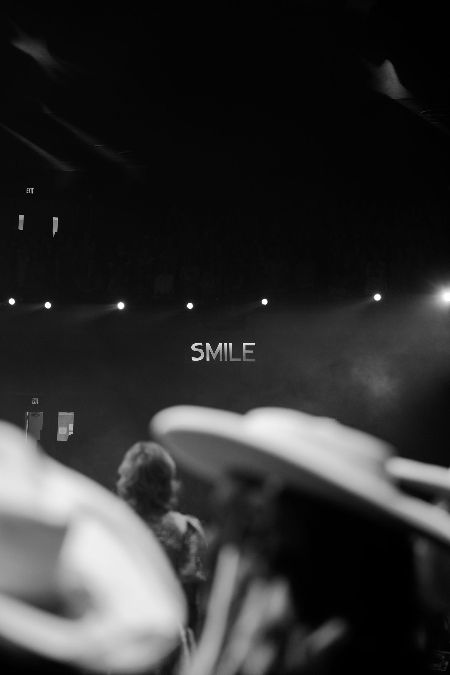 Black and white concert photo with blurred cymbals in foreground and illuminated SMILE sign on dark stage backdrop.