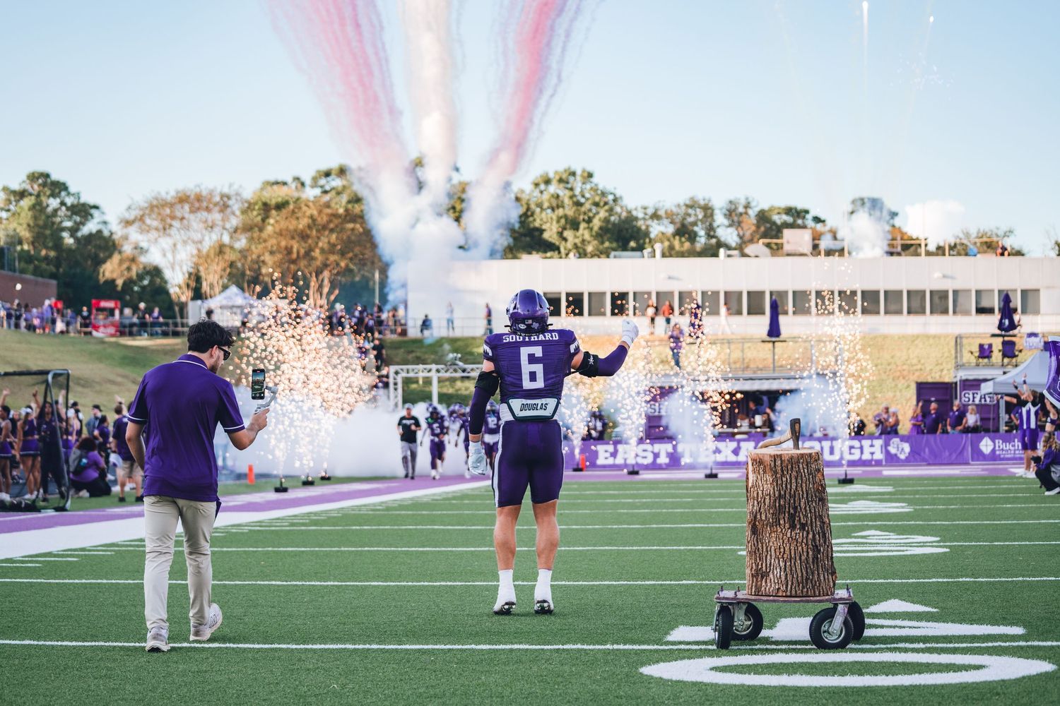 Football players and spectators gather on a field during a sports event at sunset with purple lighting effects.