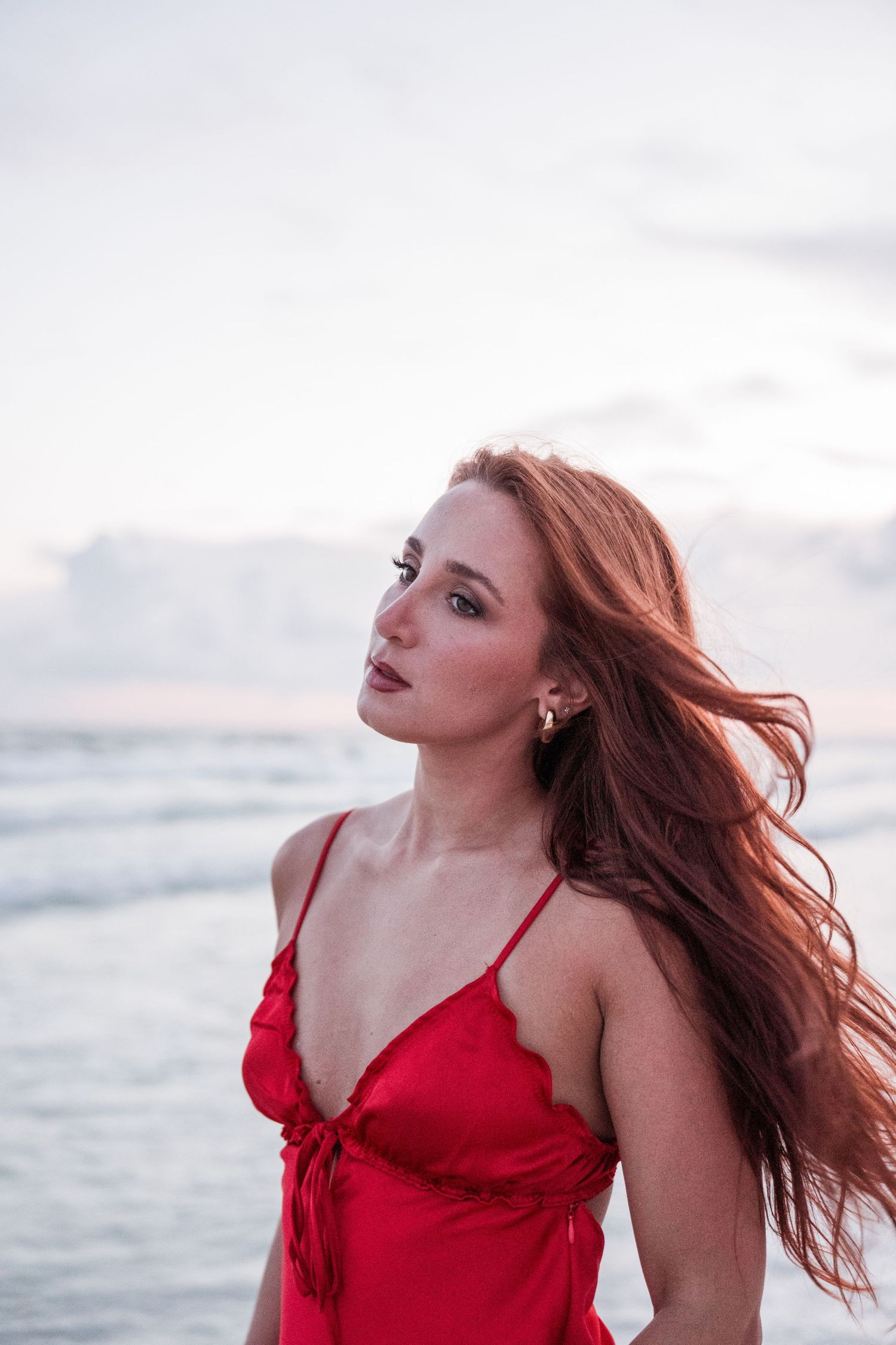 Woman in red dress with windswept hair poses by the ocean at sunset.