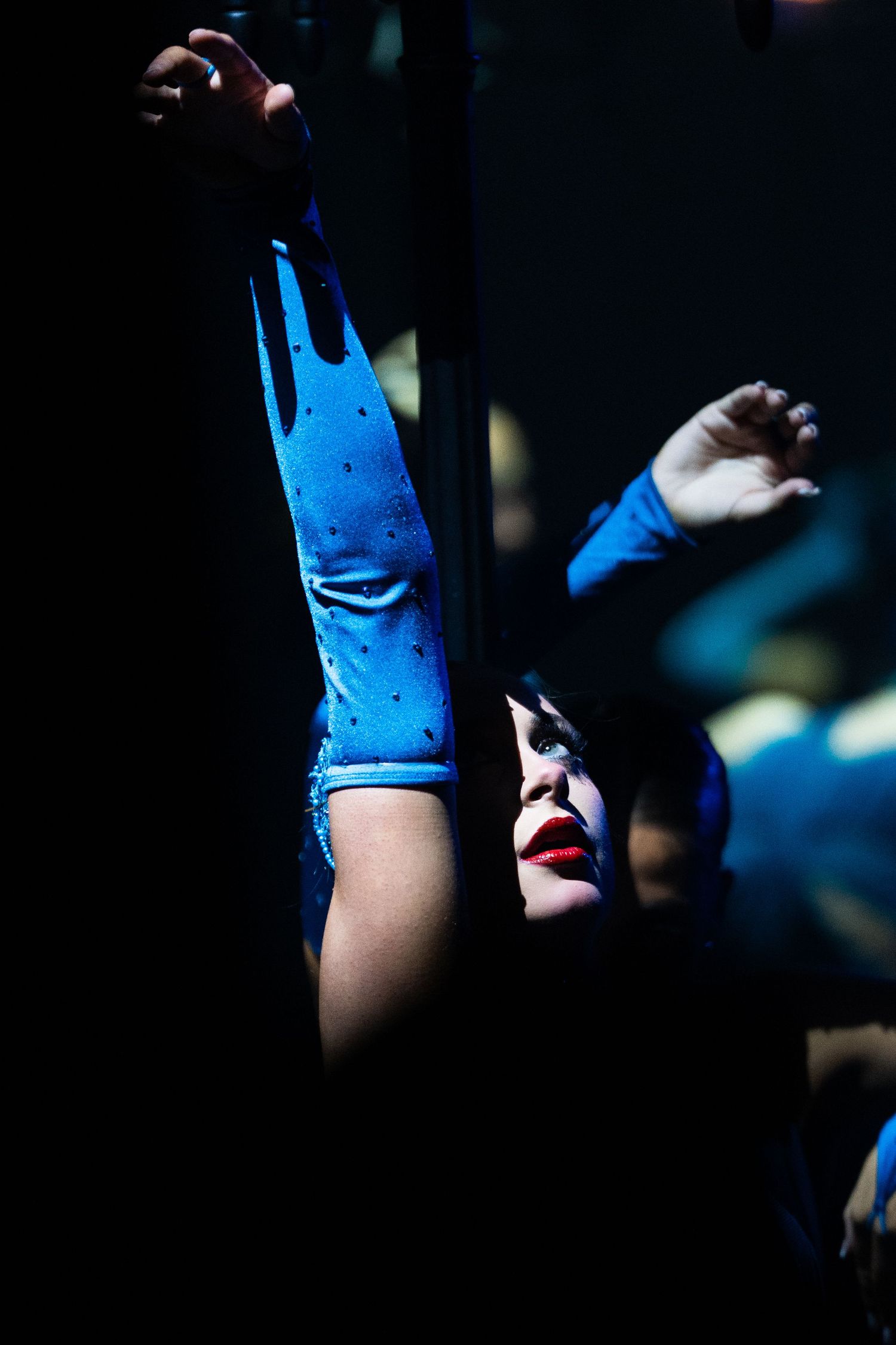 Performer in blue gloves reaching up dramatically under stage lighting with bold red lips.
