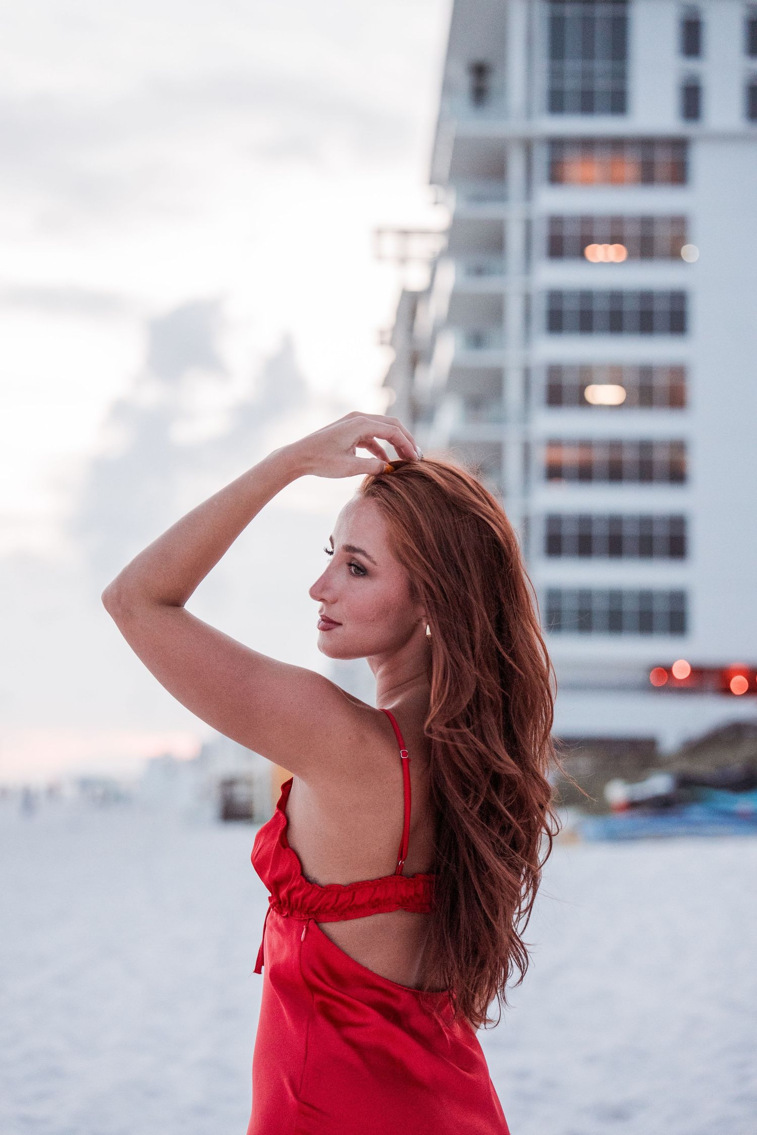 Woman in red dress poses against beachfront high-rise building during sunset.