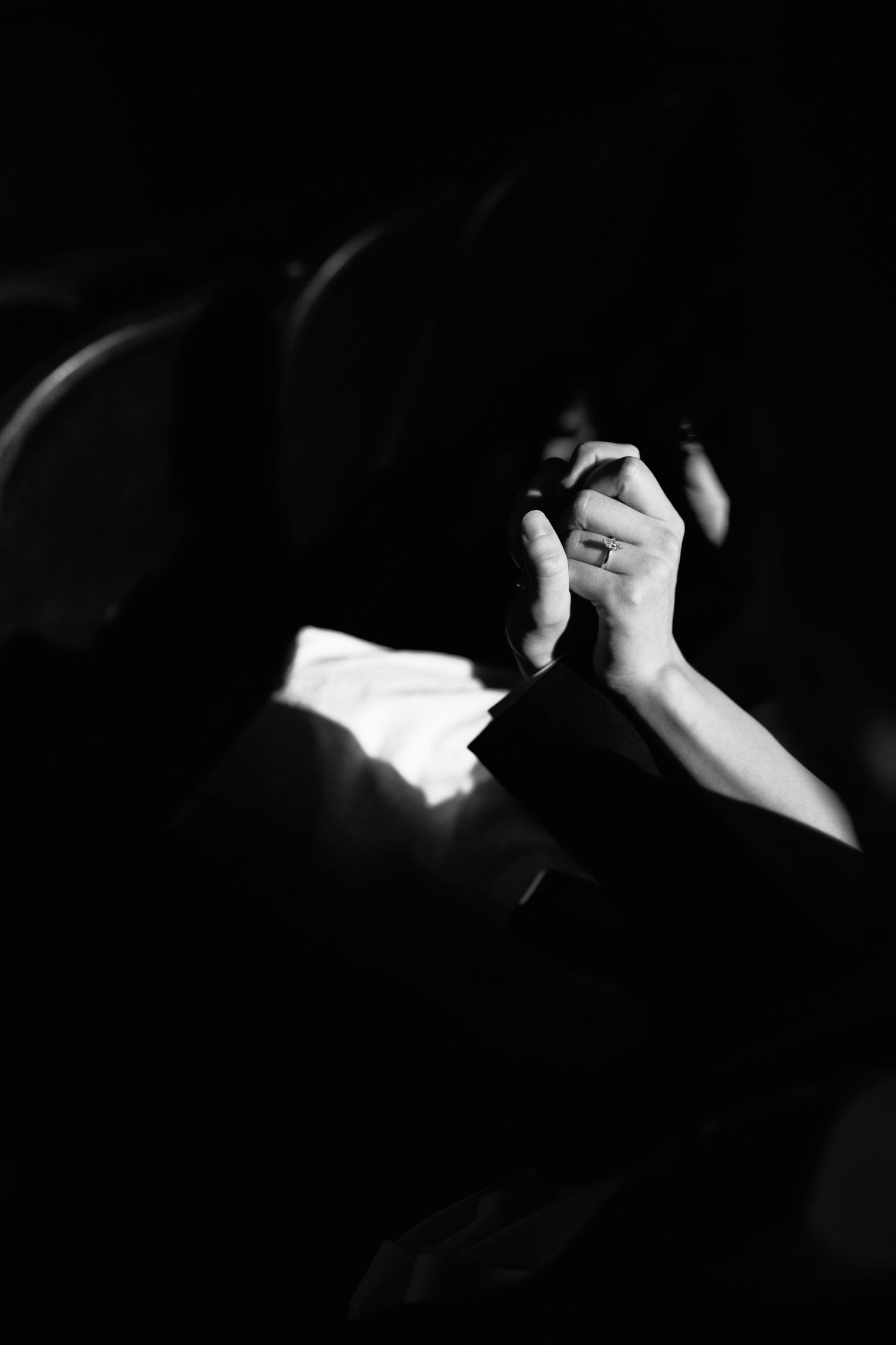 Black and white photo of clasped hands with a ring, illuminated against a dark background.