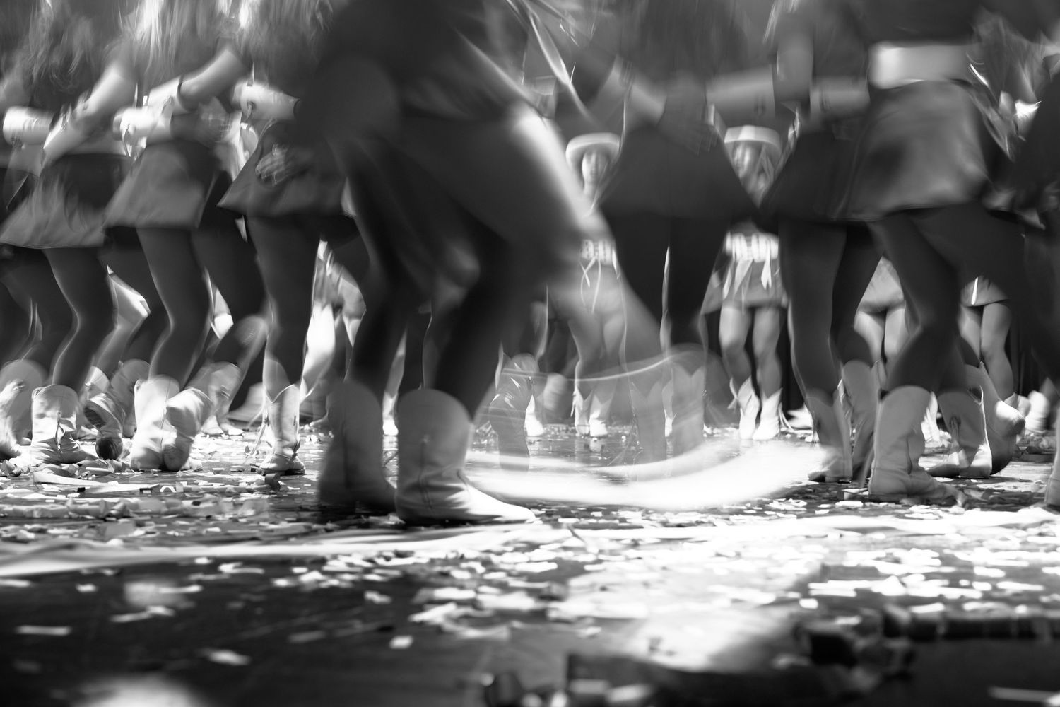 Black and white photo of dancers legs in motion on a confetti-covered floor during a festive parade.