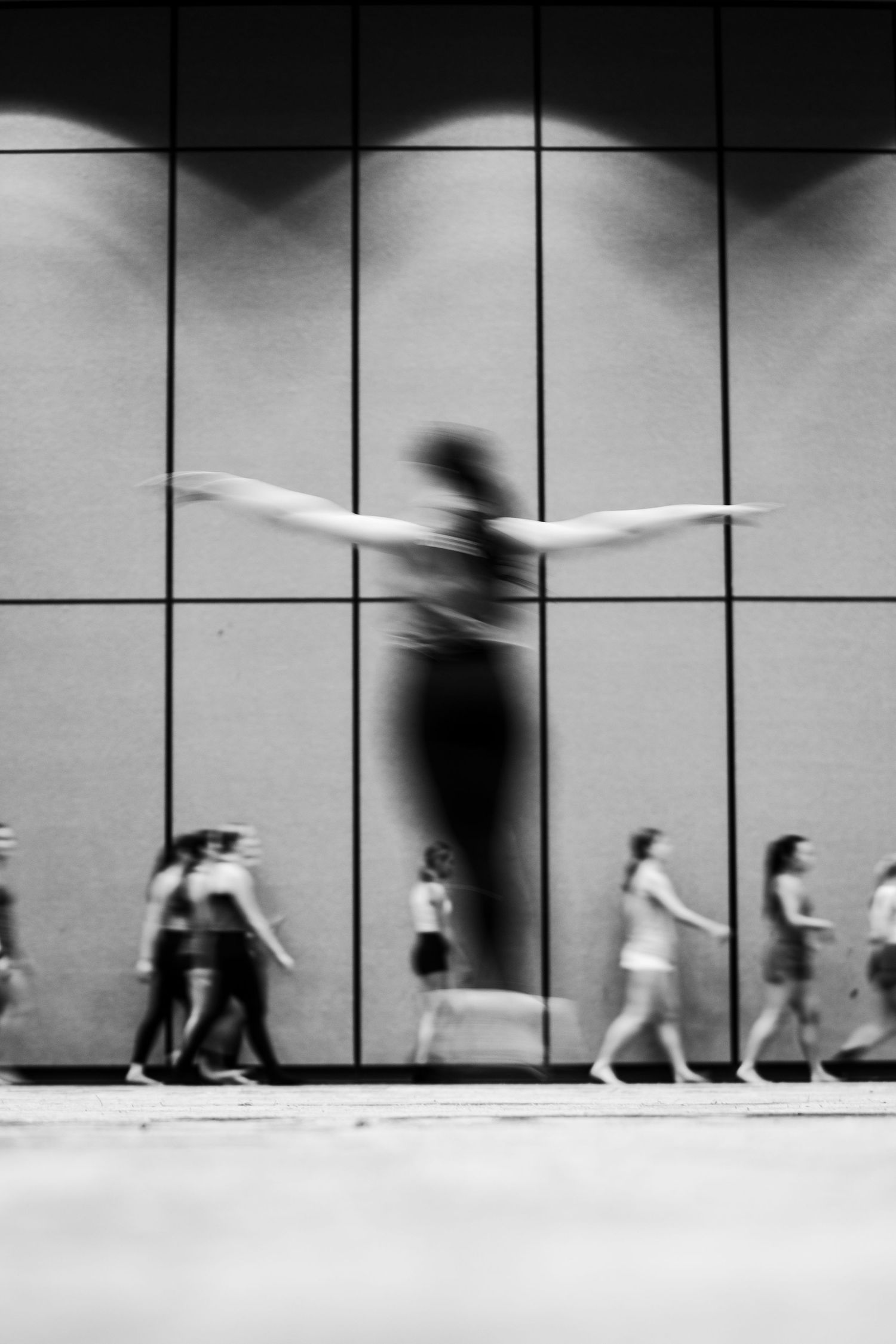 Black and white photo of a dancer with outstretched arms projected on a wall above blurred pedestrians walking by.