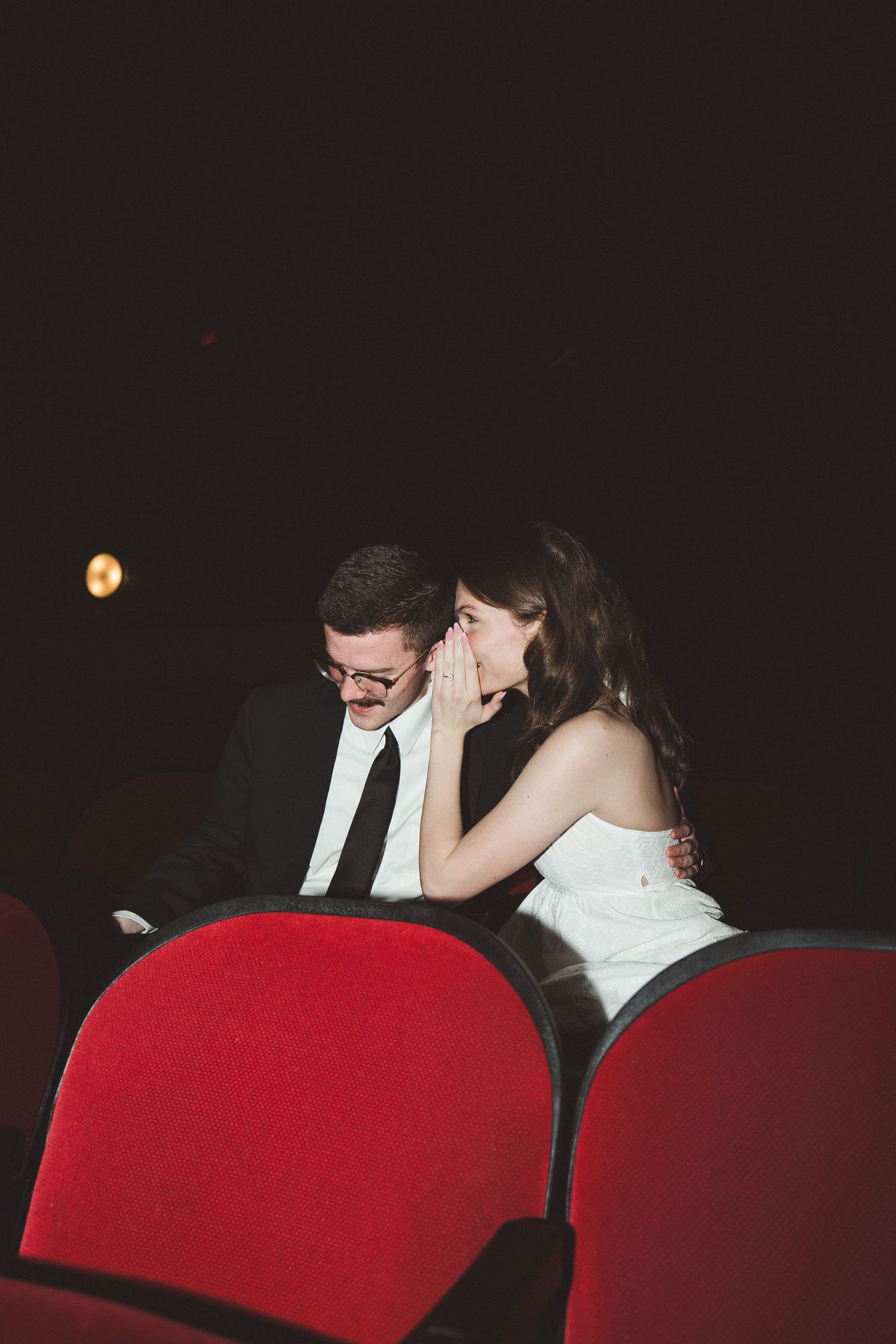 A bride whispers to her groom while sitting in red theater seats during their wedding reception.