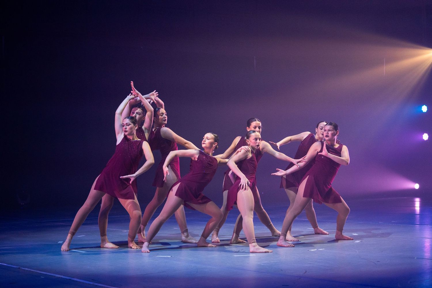 Group of female dancers in burgundy costumes performing a synchronized contemporary dance routine on a blue-lit stage.