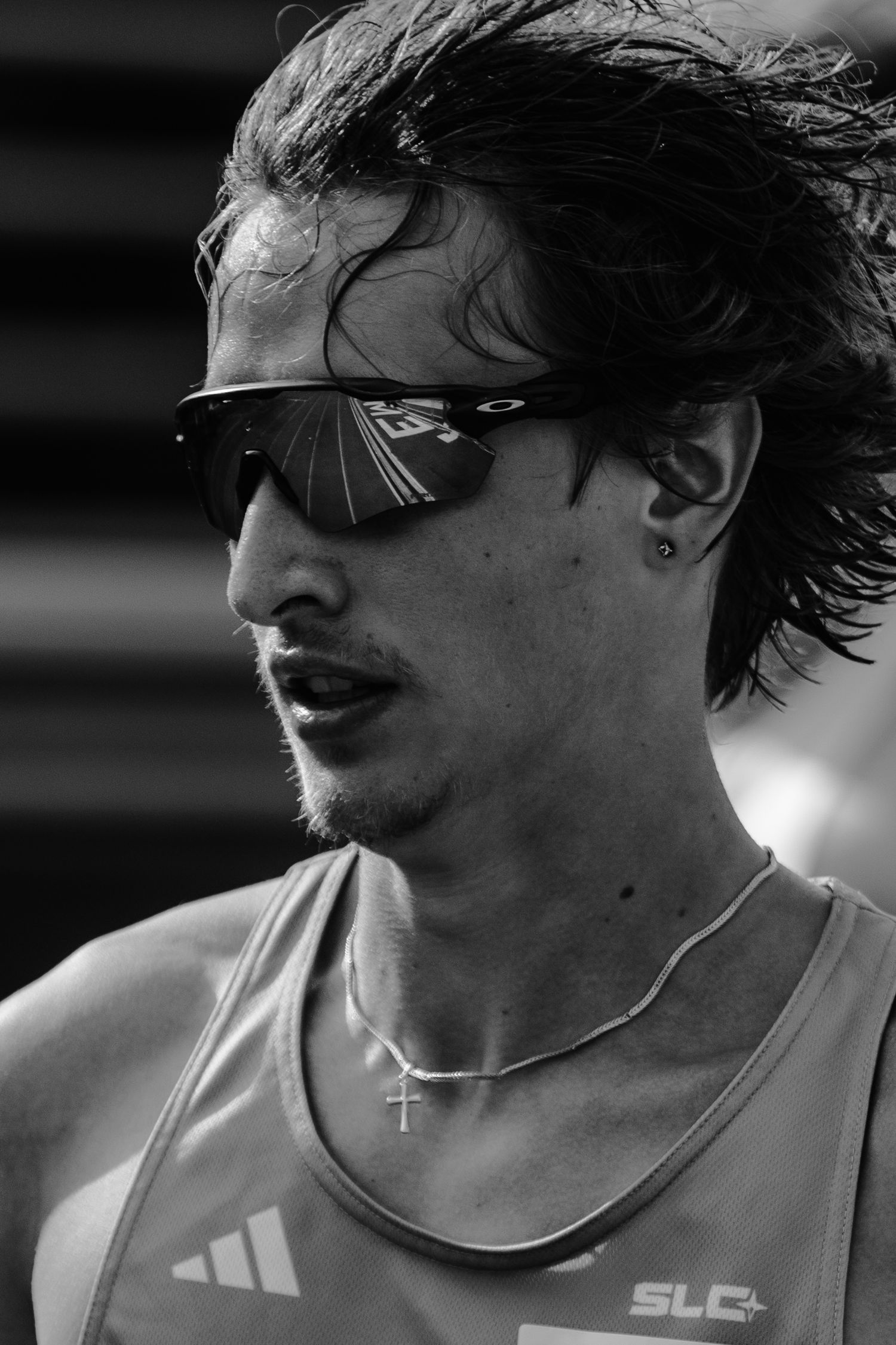 Black and white photo of a male athlete running, wearing Oakley sunglasses, Adidas jersey, cross necklace, and ear stud.