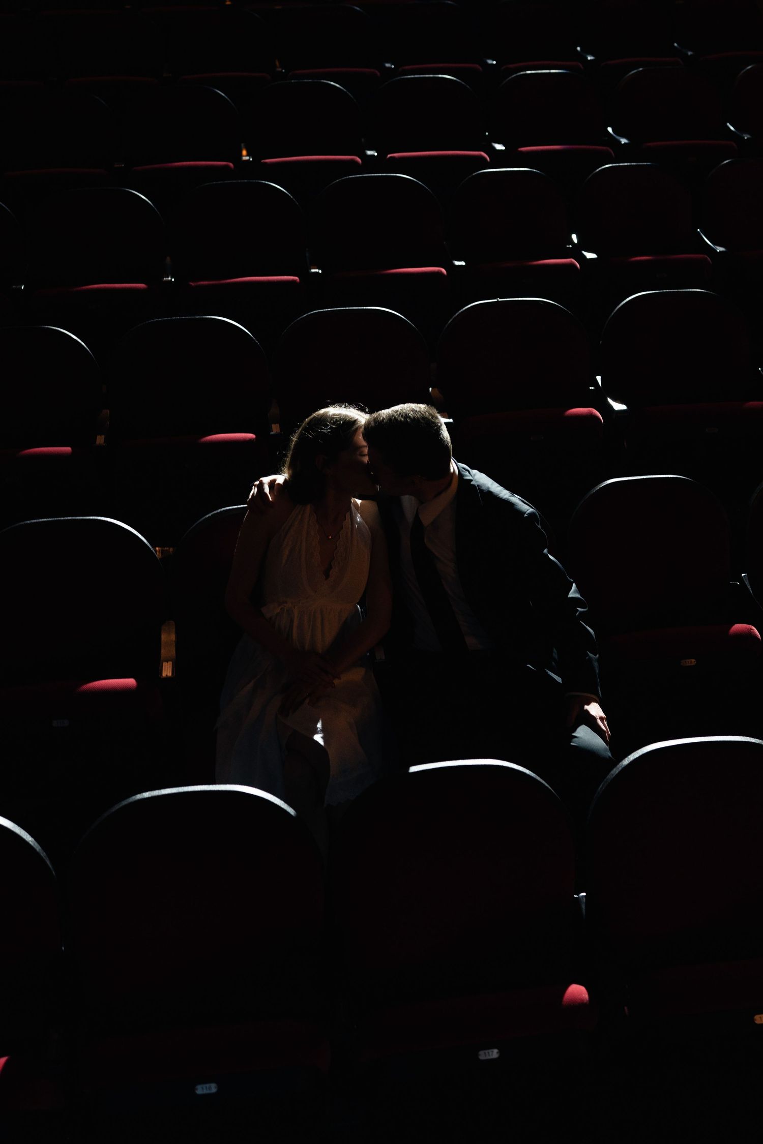 Couple sharing a romantic moment in a dimly lit empty theater with rows of red and black seats.