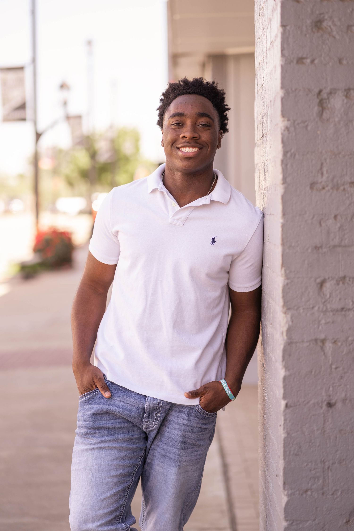 Young person in white polo shirt and jeans smiling while leaning against white wall outdoors.