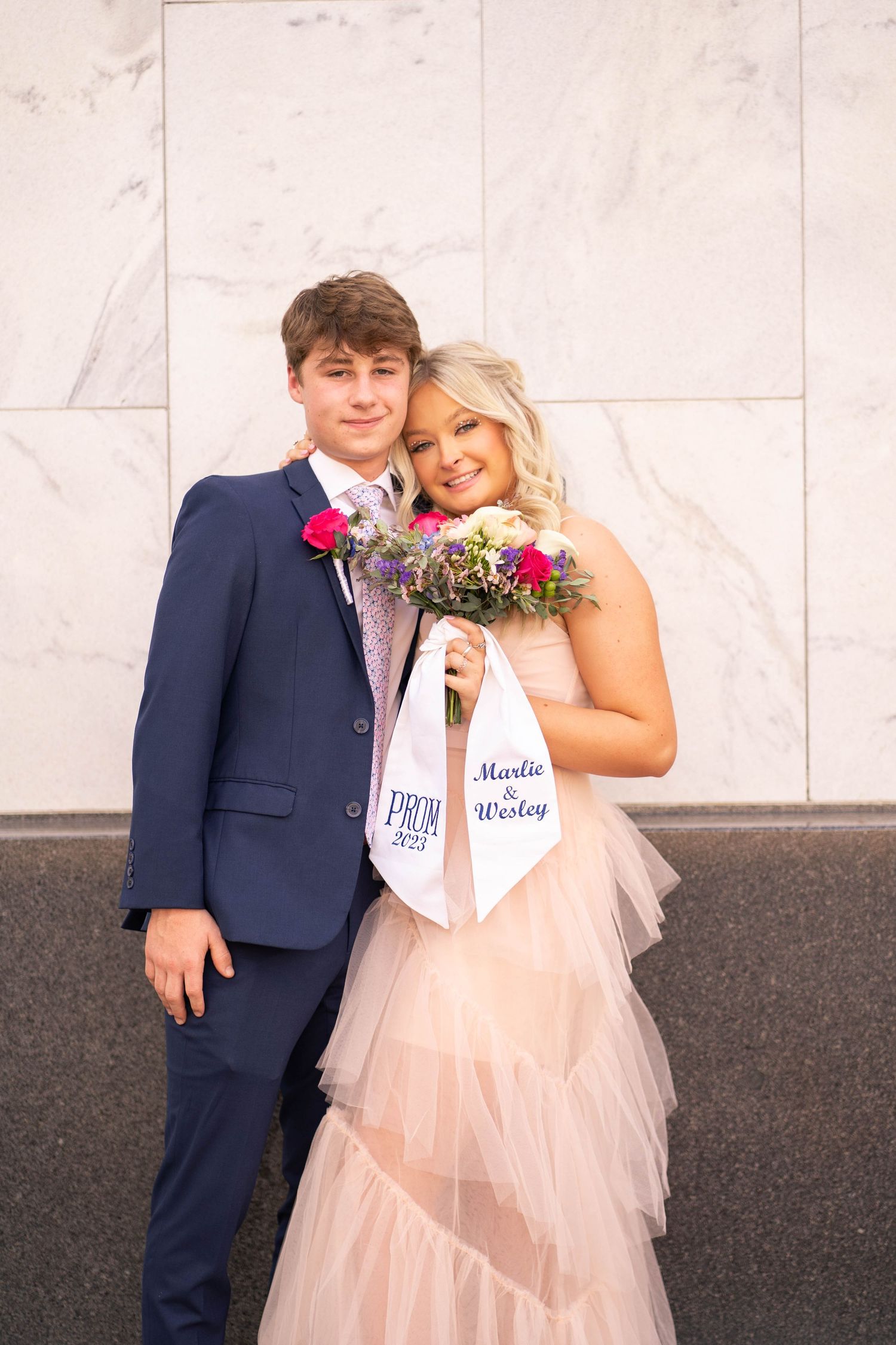 Couple poses for prom photo with boutonniere and corsage against marble wall.