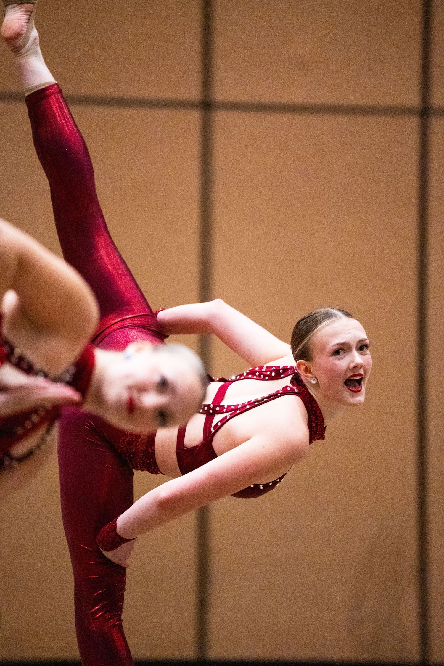 Young dancers in red sparkly costumes perform acrobatic dance routine, with one dancer doing a high leg extension.