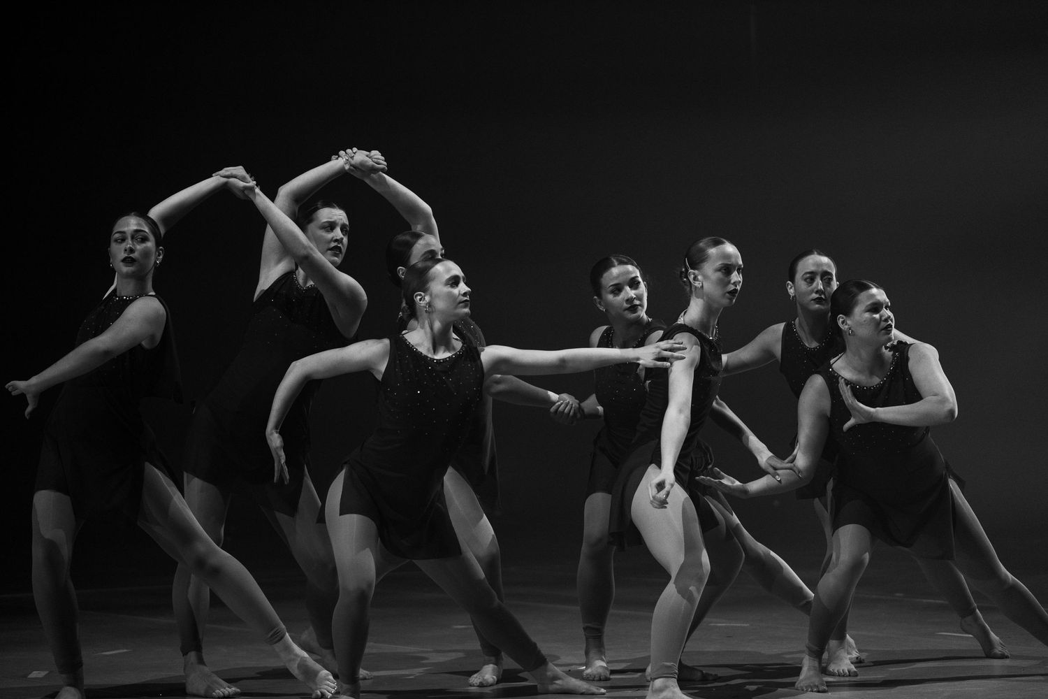 Group of dancers in black leotards performing a dramatic contemporary dance routine on a dark stage.