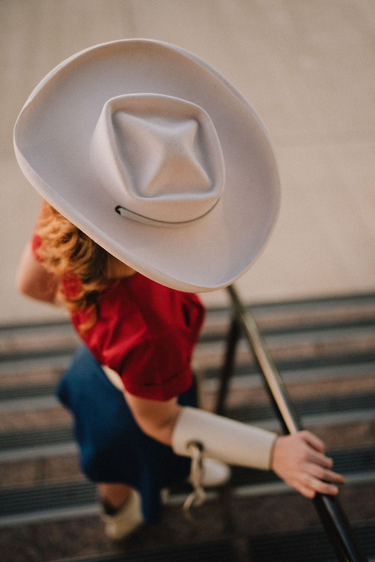 A white cowboy hat sits atop someone wearing a red shirt and blue jeans while crouching on metal bleachers.