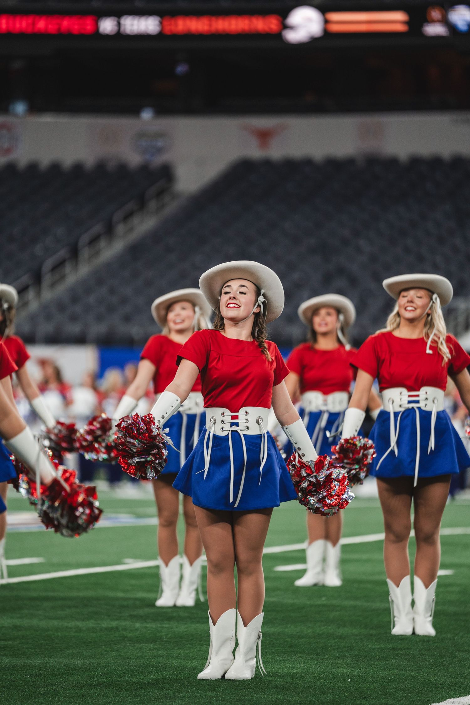 Cheerleaders in red and white uniforms with cowboy boots lined up on a football field performing synchronized routine.