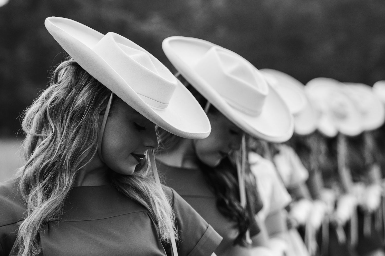 Black and white photo of women in wide-brim white hats standing in a line, heads bowed elegantly.
