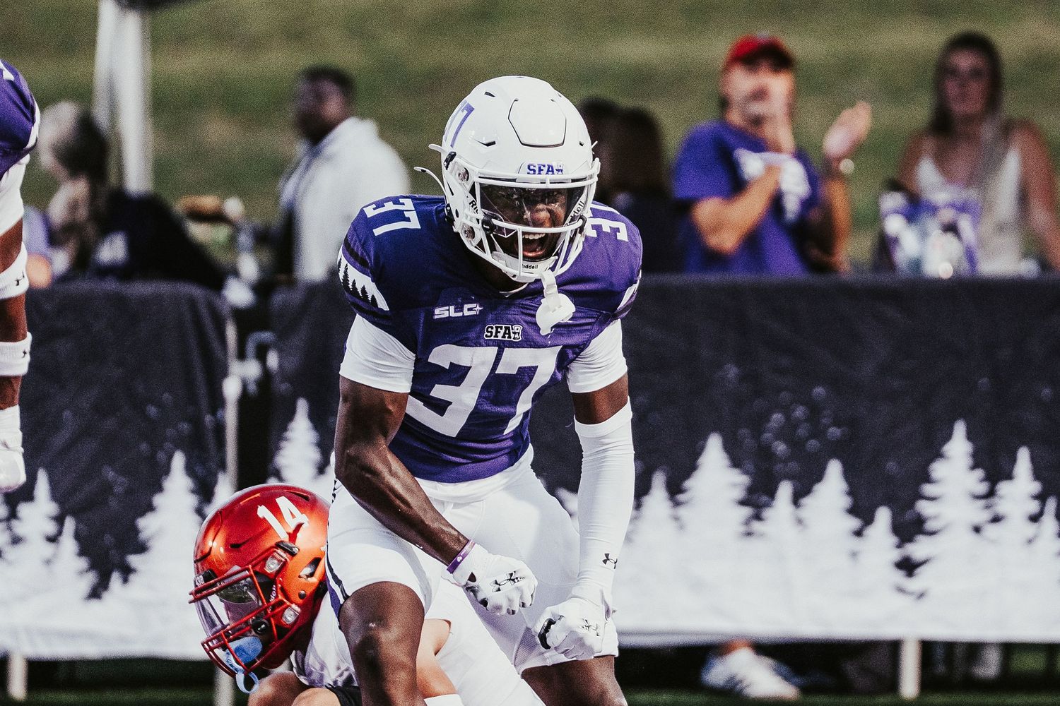 A football player in a white and purple uniform stands on the field during a game against a red-uniformed opponent.