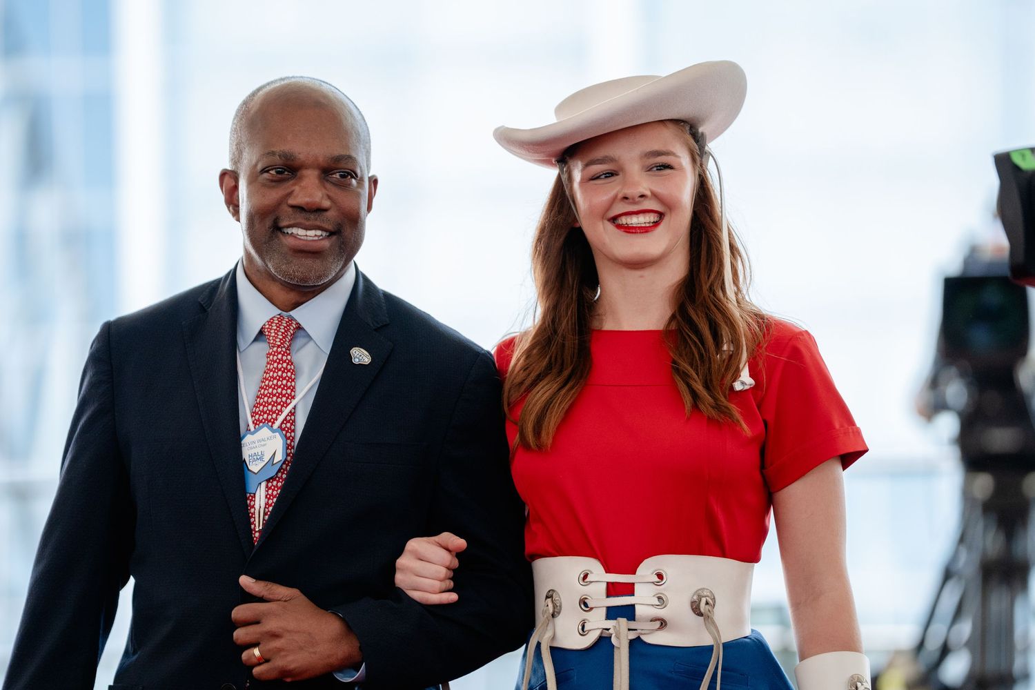 Two people walking arm in arm outdoors, one wearing a business suit and the other in a red top with a beige hat.