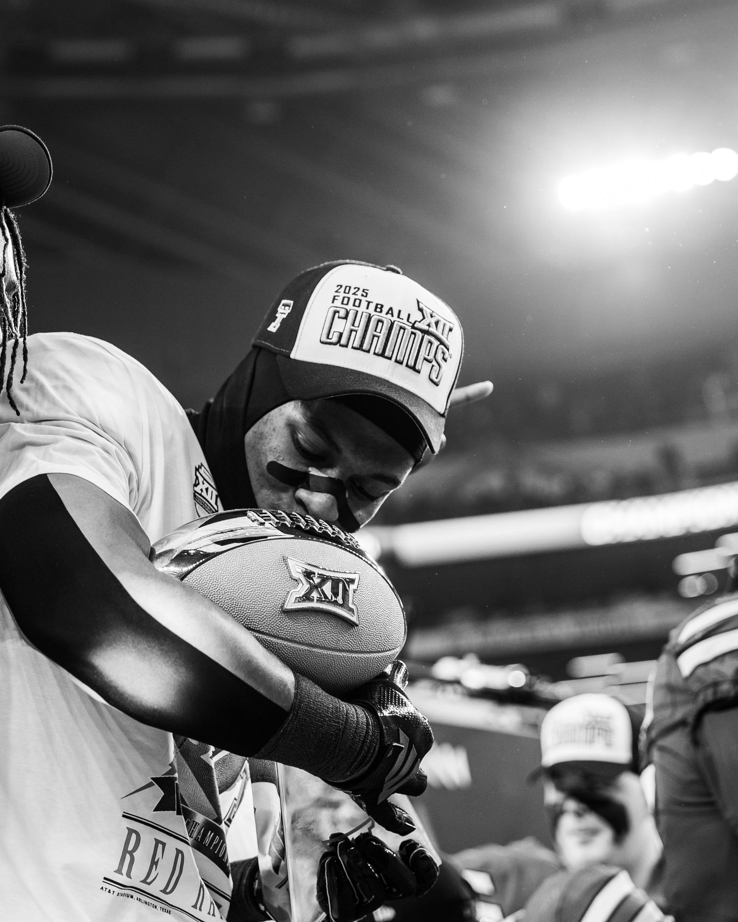 Black and white photo of a football player holding a Big 12 ball wearing a 2025 Football Champions hat.