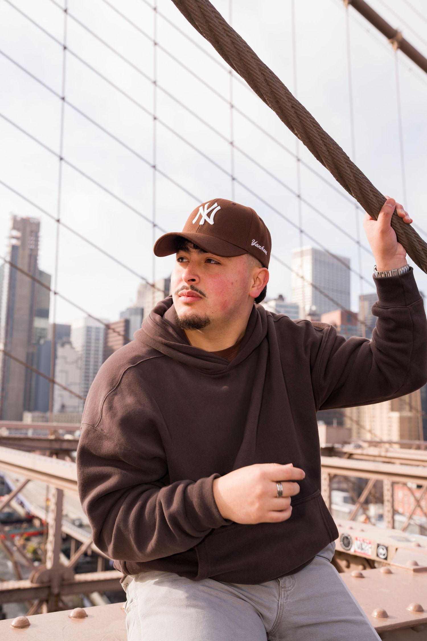Person in brown hoodie and Yankees cap posing on Brooklyn Bridge with NYC skyline in background.