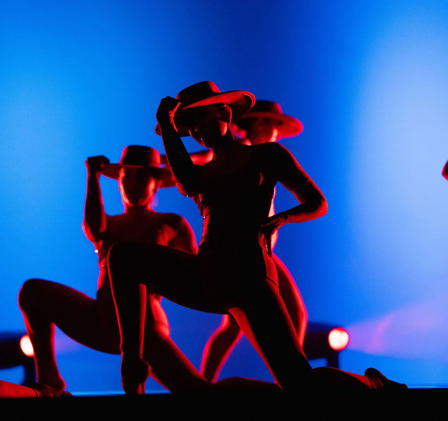 Silhouetted dancers in dramatic red and blue lighting strike powerful poses on stage wearing cowboy hats.
