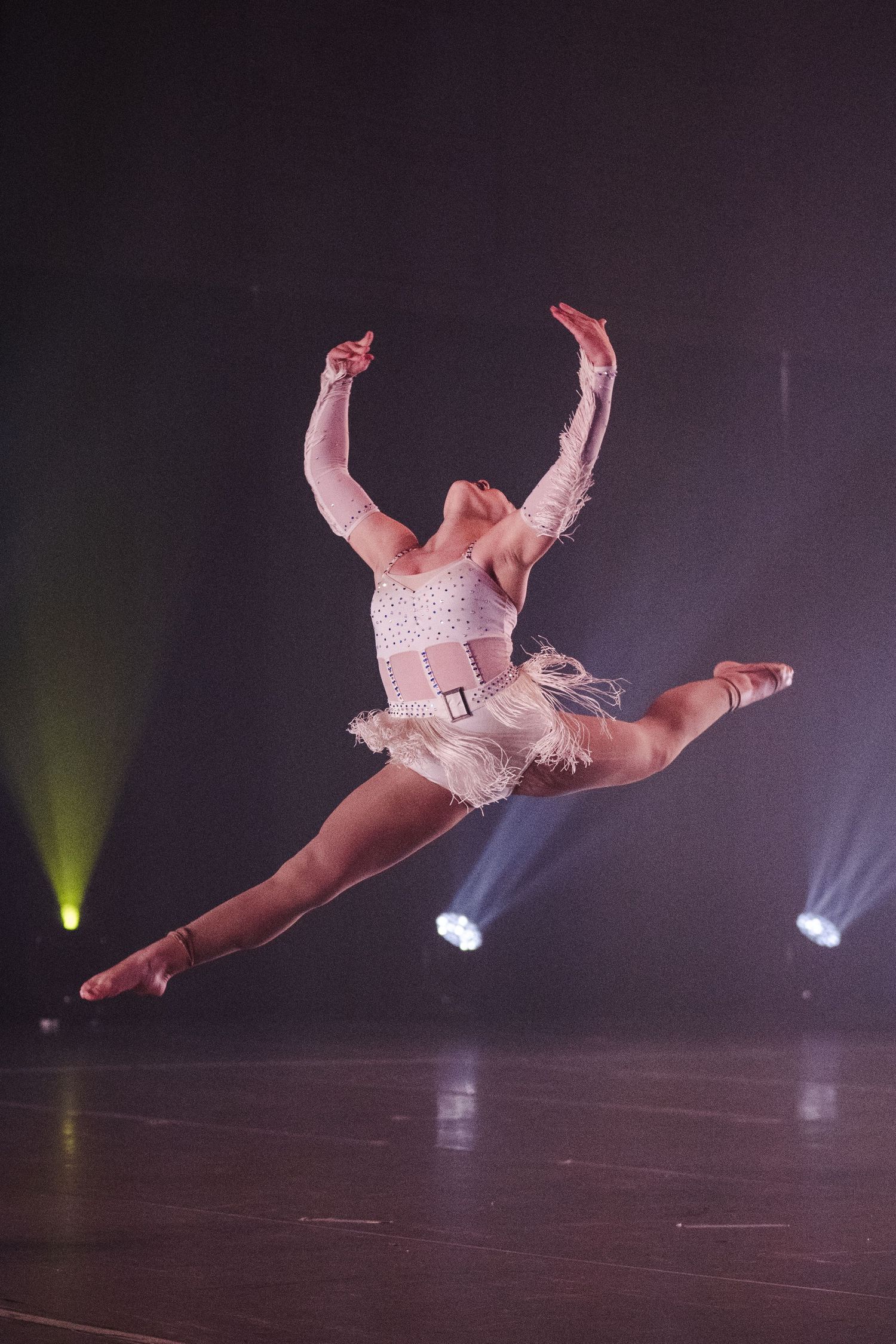Dancer leaping mid-air in white feathered costume on dark stage with dramatic green and blue spotlights.