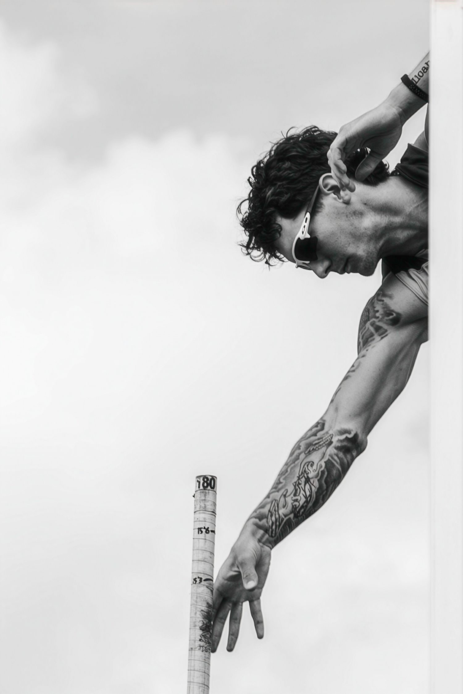 Black and white photo of a tattooed shirtless man with sunglasses leaning over a ledge against a cloudy sky.