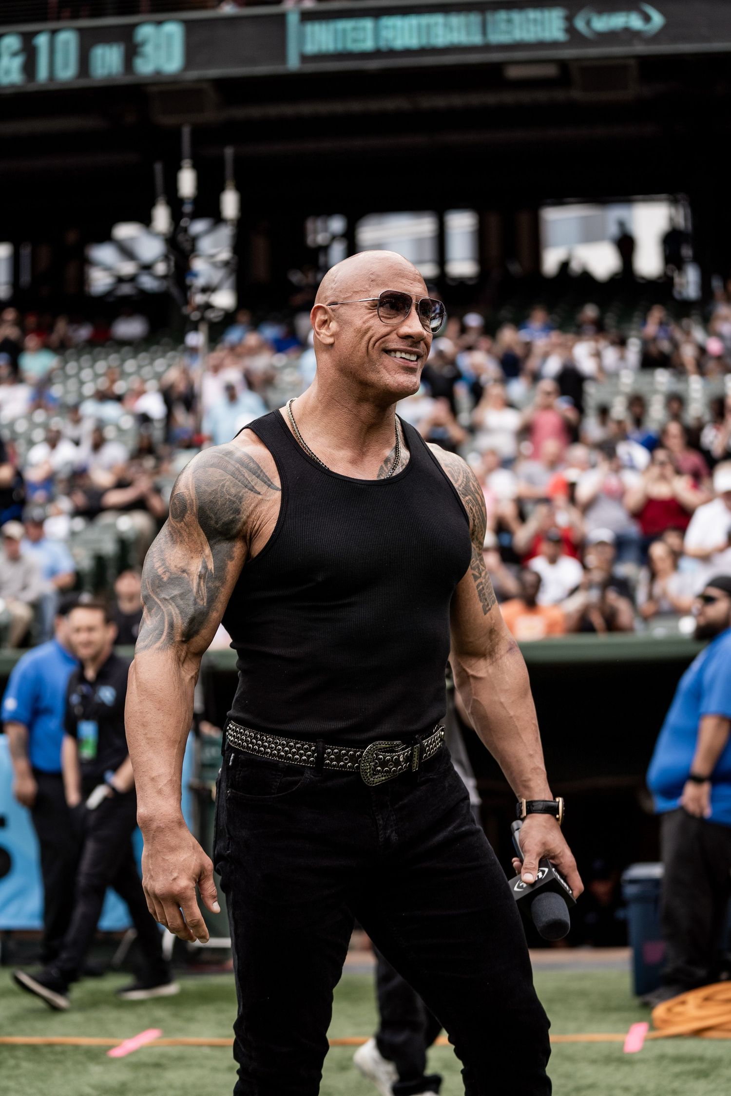 Athletes in black tank tops compete during an outdoor sporting event with crowd in background.