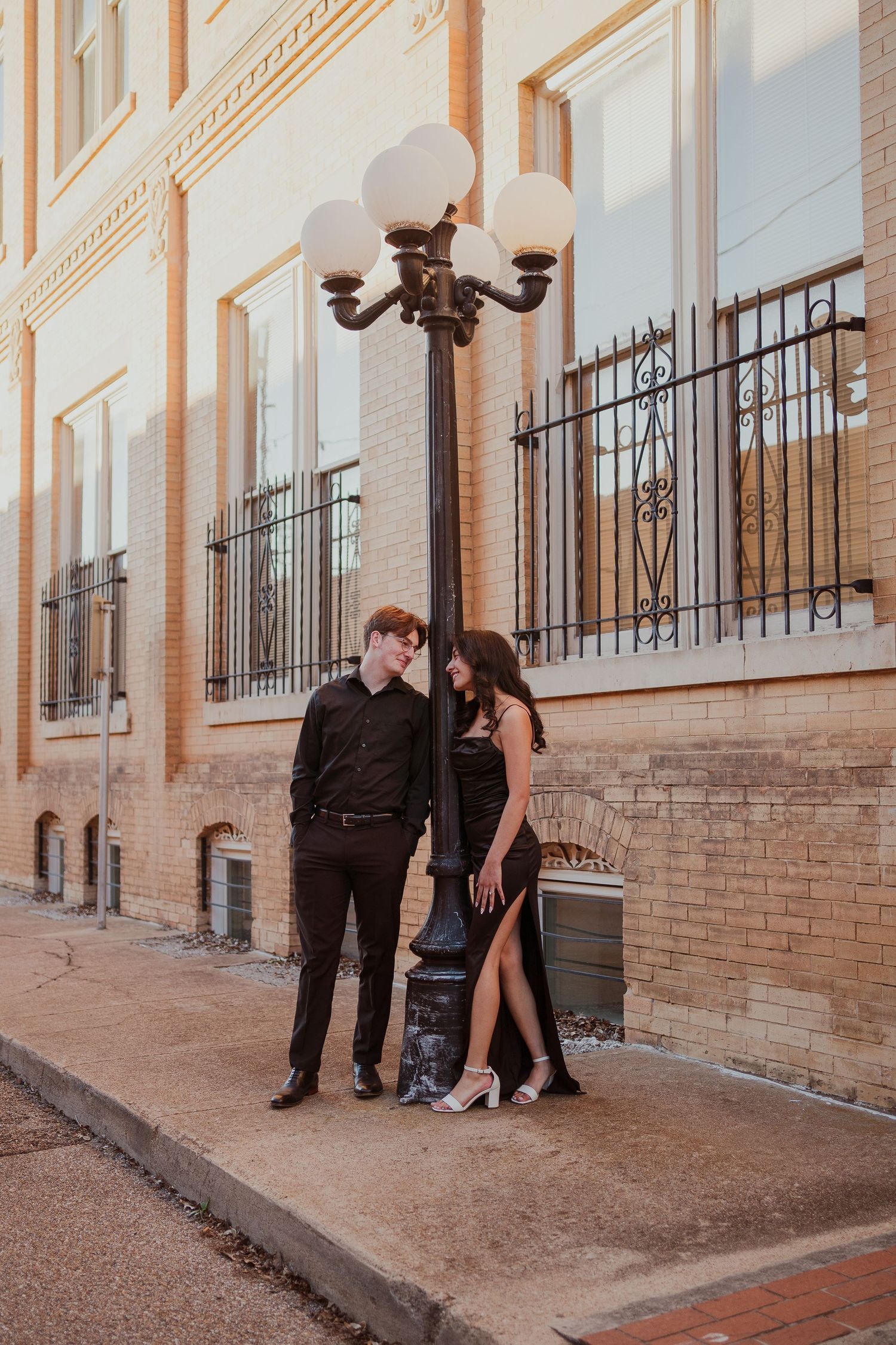 Couple poses under vintage street lamp against brick building at dusk.