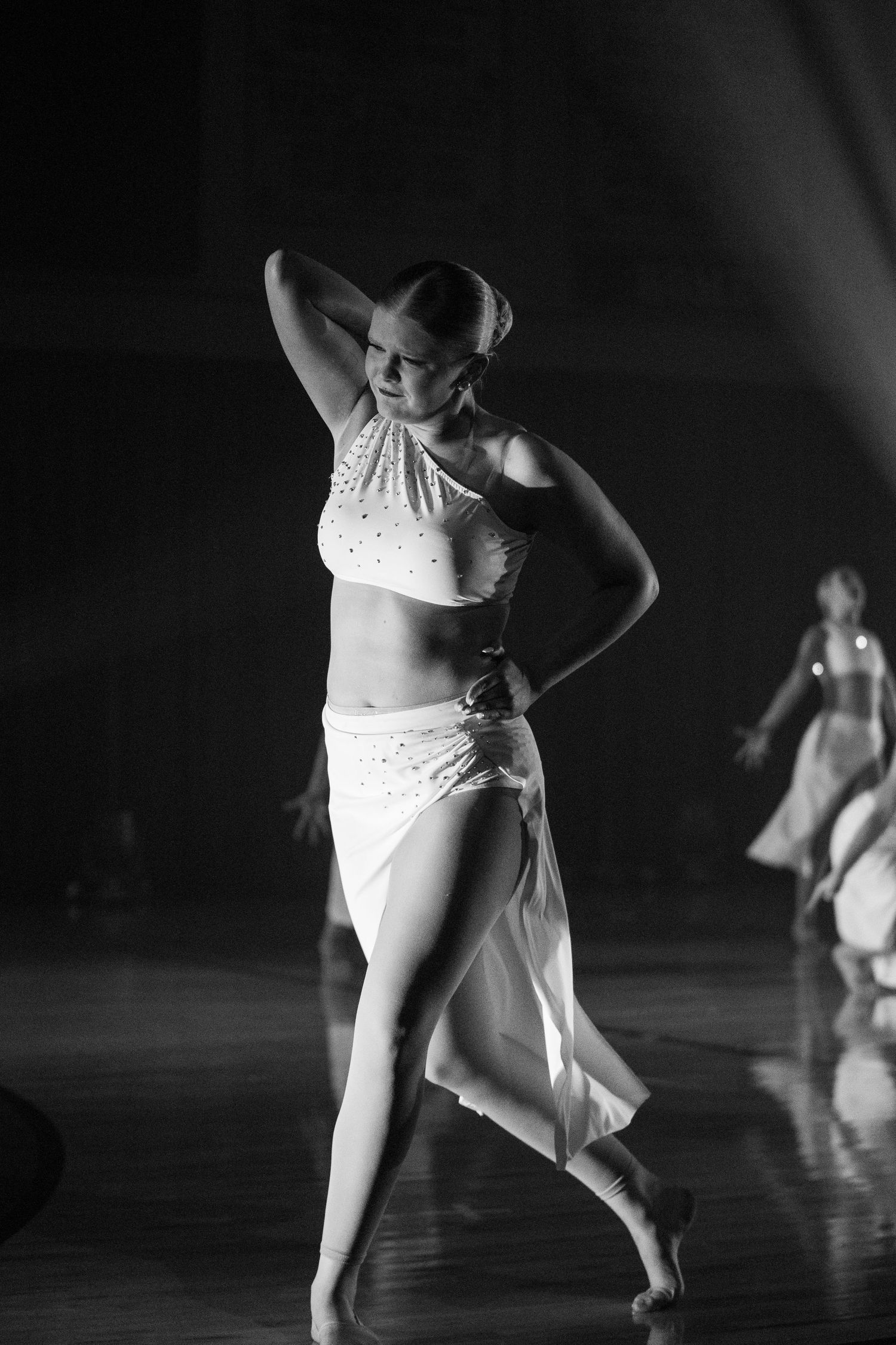Black and white photo of a dancer in white costume performing a dynamic movement on a dark stage.
