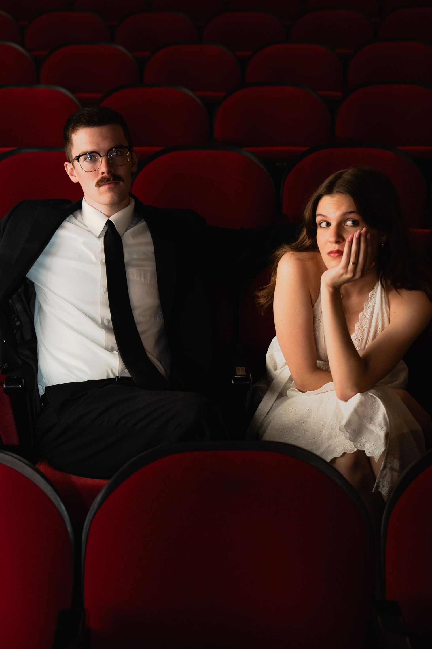Couple in formal attire sitting in red theater seats, man in suit with mustache, woman in white dress looking pensive.